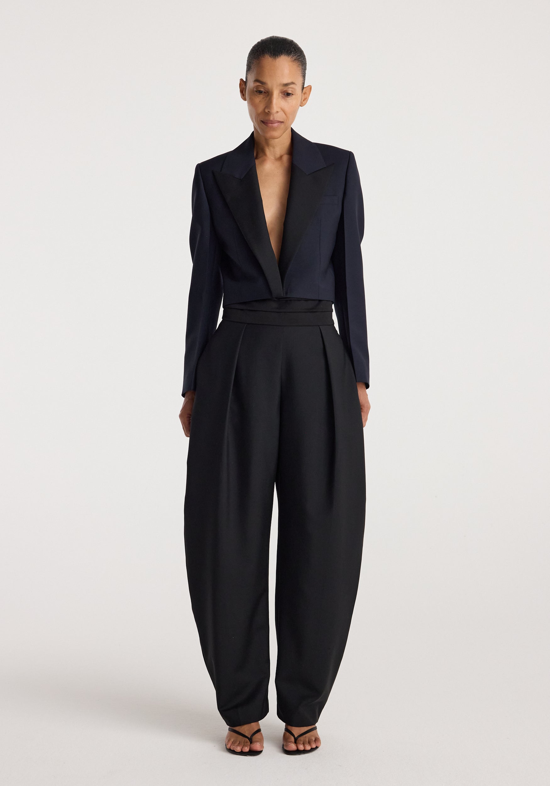 Woman wearing a tailored Cropped blazer | navy with long sleeves and wide lapels paired with high-waisted black trousers and open-toe heels, standing against a plain background.