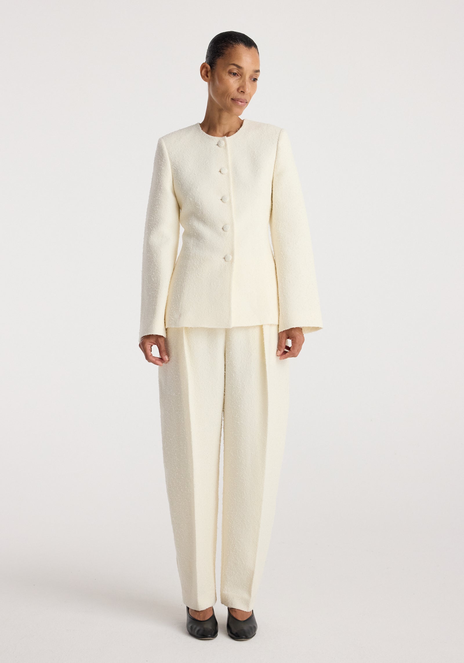 Woman wearing a cream textured bouclé shaped blazer | cream with matching wide-legged pants, black shoes, standing against a plain white background.