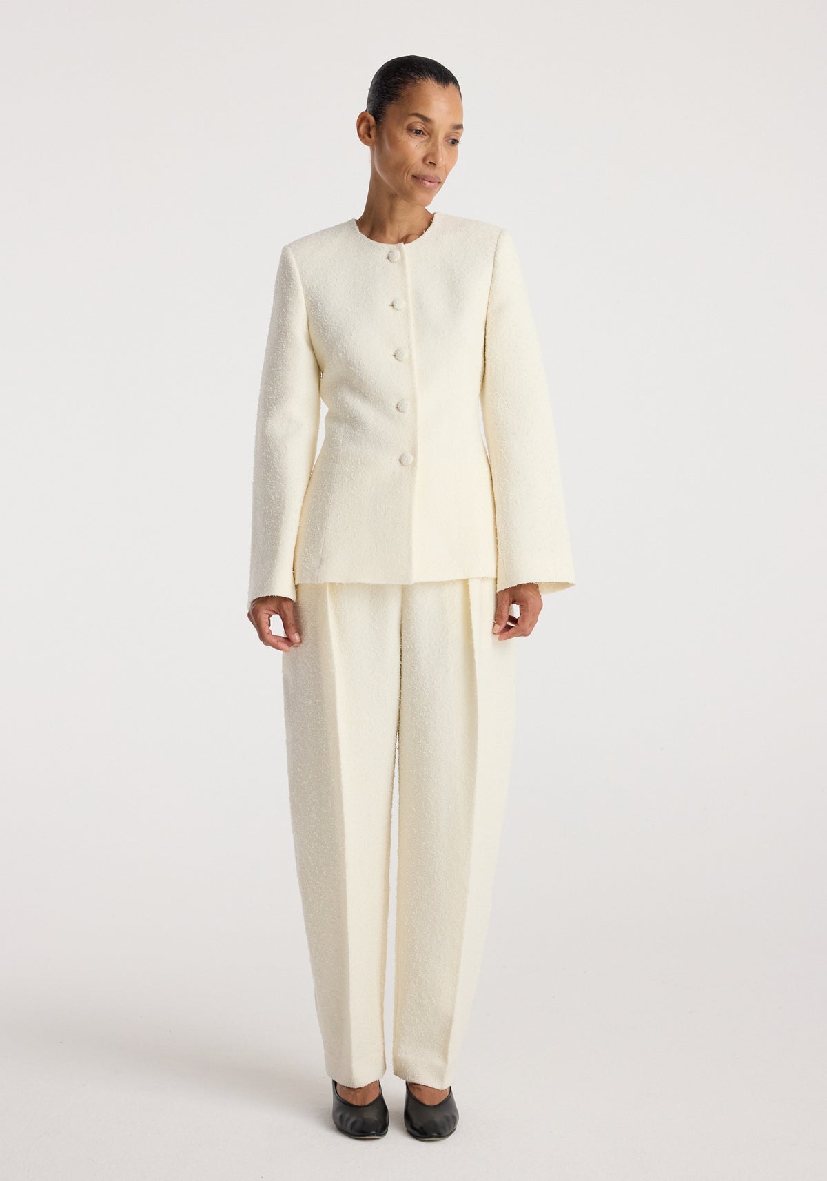 Woman wearing a cream textured bouclé shaped blazer | cream with matching wide-legged pants, black shoes, standing against a plain white background.