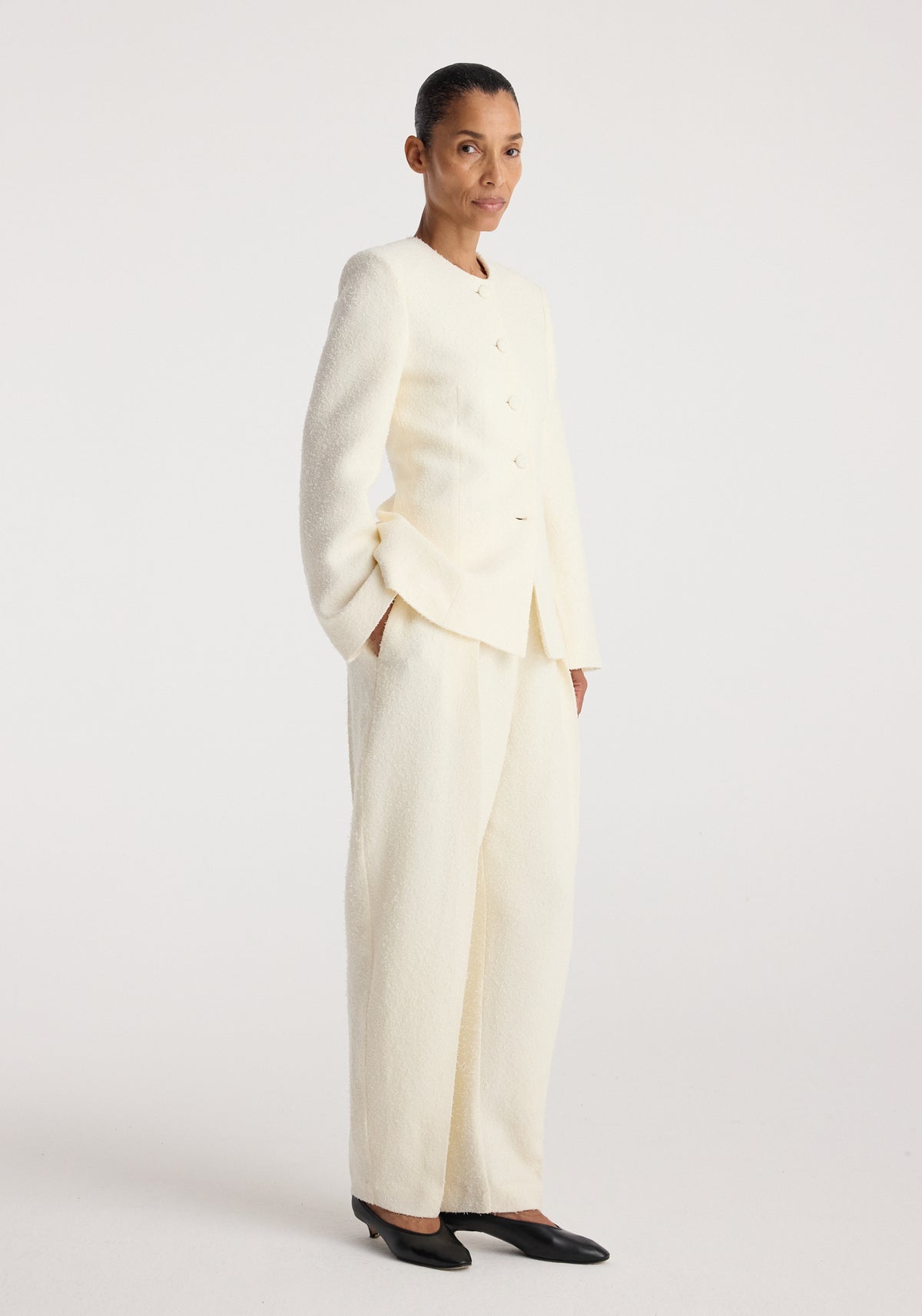 Woman wearing a cream-colored bouclé shaped blazer | cream with matching wide-leg trousers and black pointed flats, standing against a plain white background.