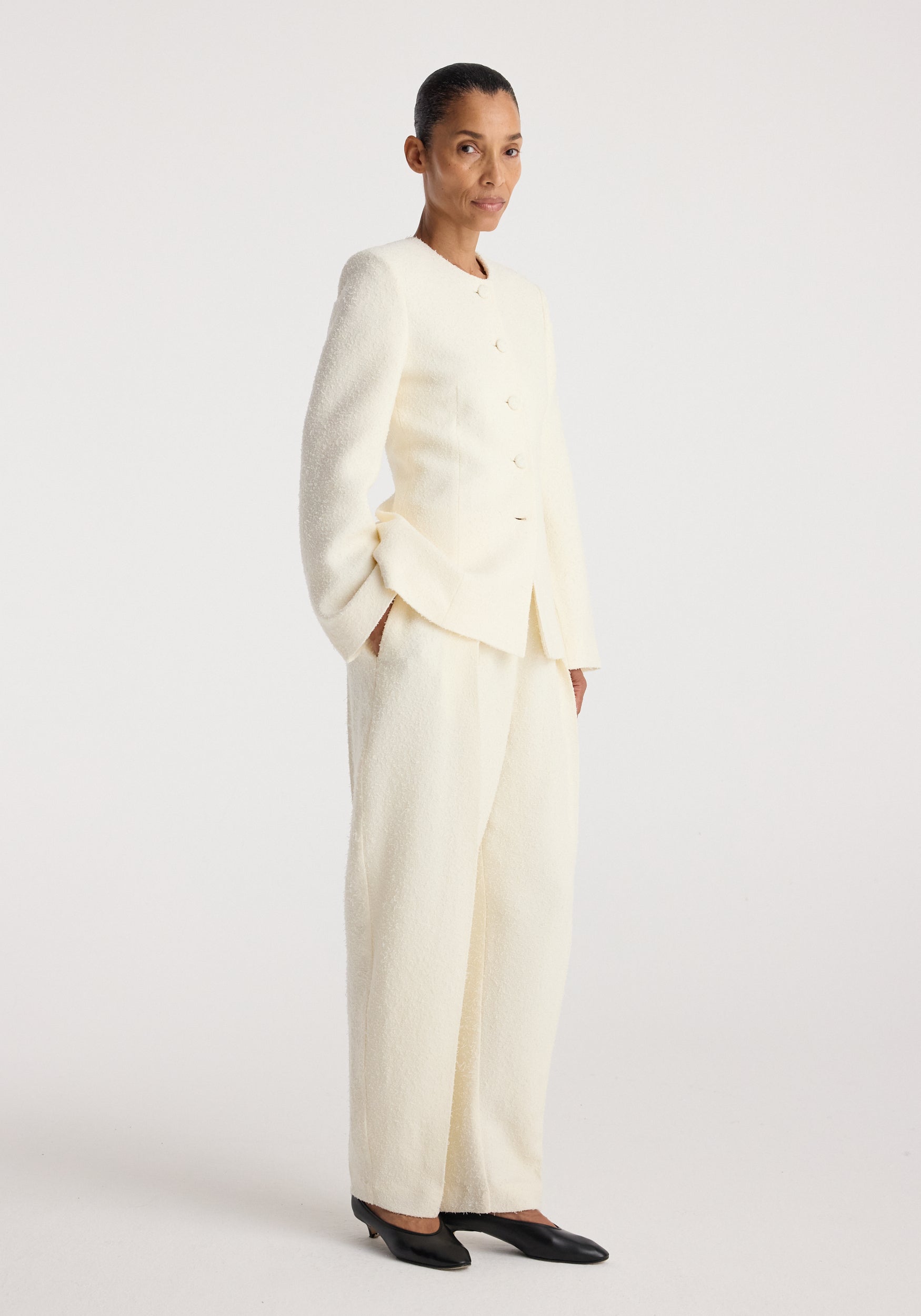 Woman wearing a cream-colored bouclé shaped blazer | cream with matching wide-leg trousers and black pointed flats, standing against a plain white background.