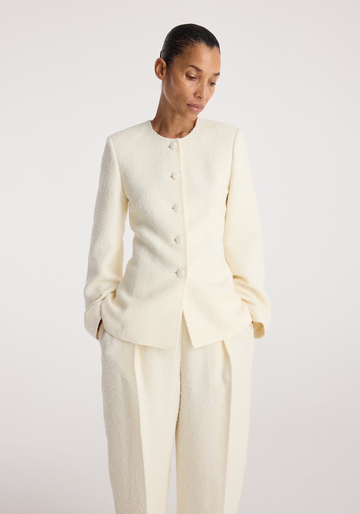 Woman wearing a cream textured long-sleeve Bouclé shaped blazer | cream with covered buttons, paired with matching trousers, standing against a plain background.