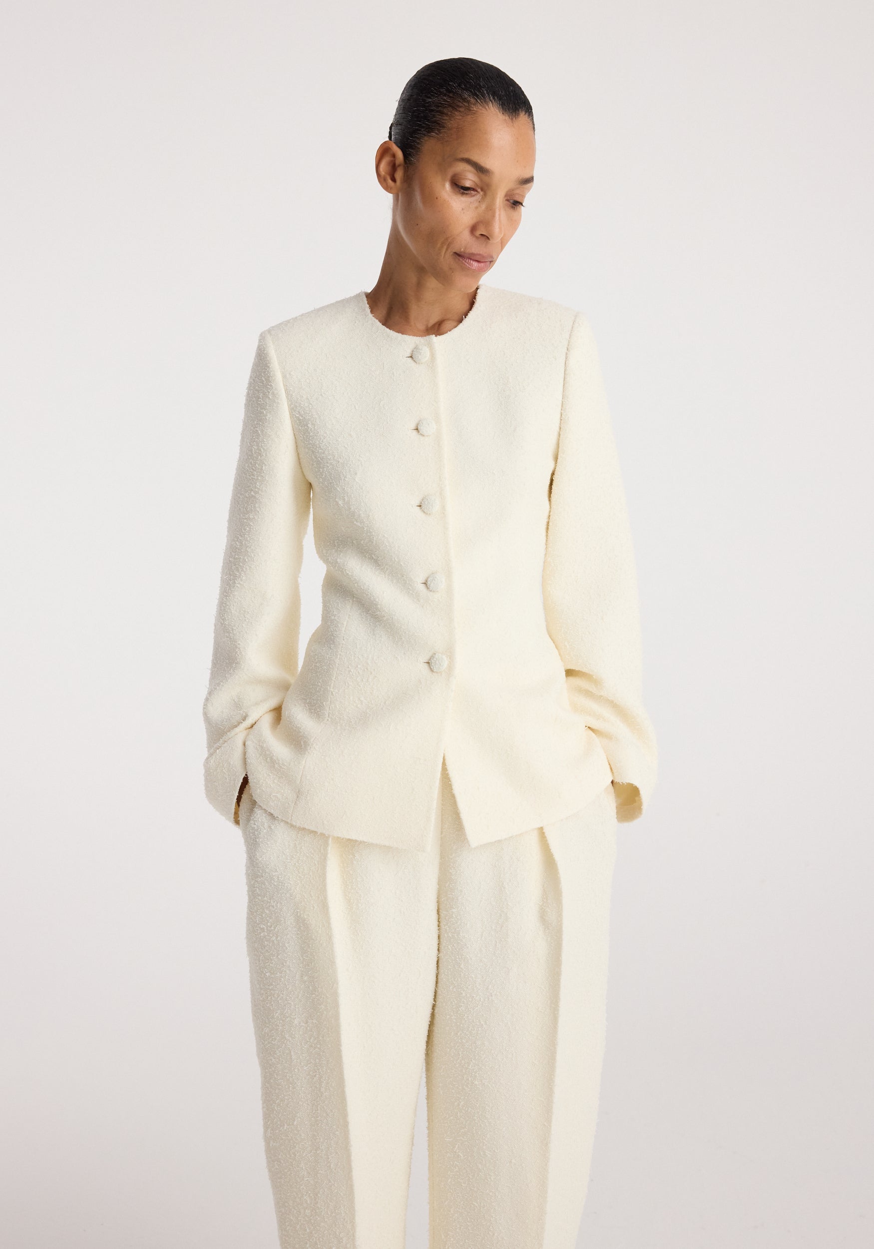 Woman wearing a cream textured long-sleeve Bouclé shaped blazer | cream with covered buttons, paired with matching trousers, standing against a plain background.