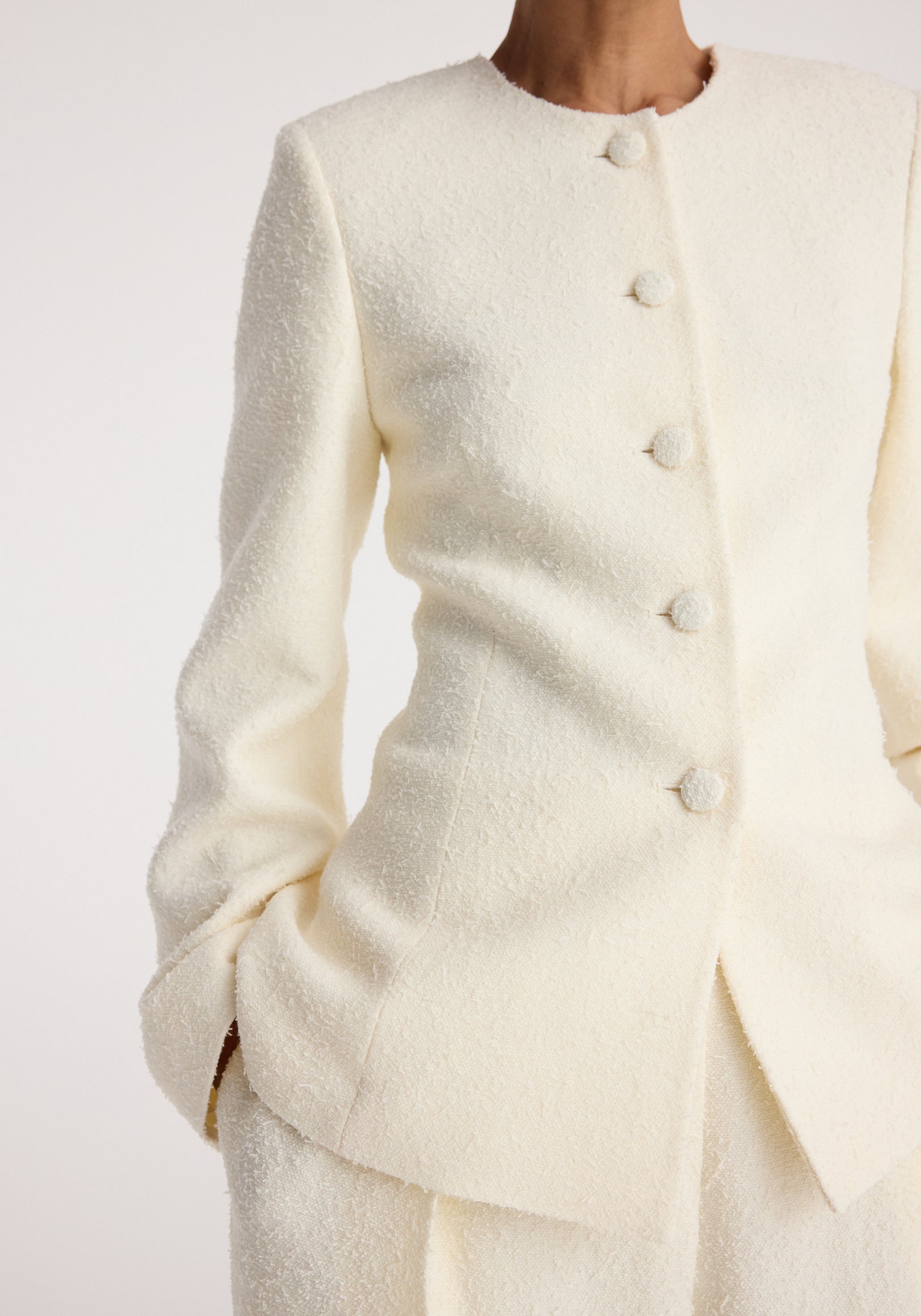 Close-up of a cream textured bouclé shaped blazer | cream with button detailing and long sleeves worn with matching pants.