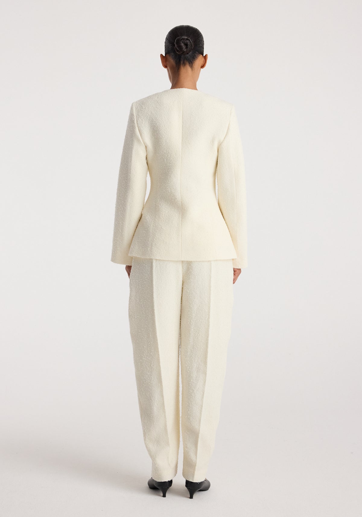 Woman wearing a cream textured bouclé shaped blazer | cream with matching wide-leg trousers, viewed from the back against a plain background.