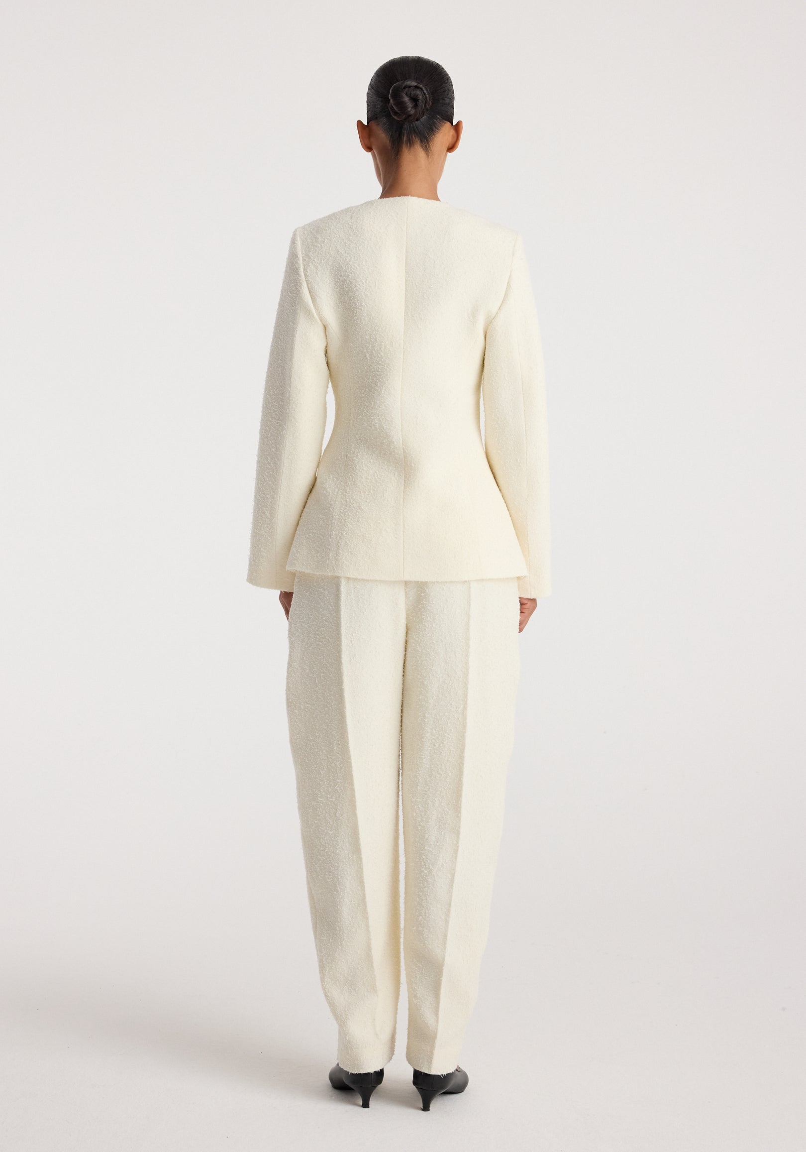 Woman wearing a cream textured bouclé shaped blazer | cream with matching wide-leg trousers, viewed from the back against a plain background.