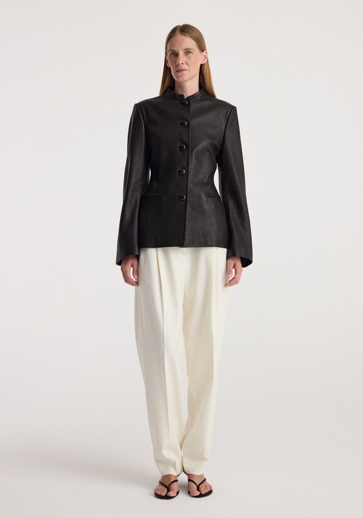 Woman wearing a tailored black shaped grain leather jacket | black with button closure, paired with loose white trousers and black sandals, standing against a plain background.