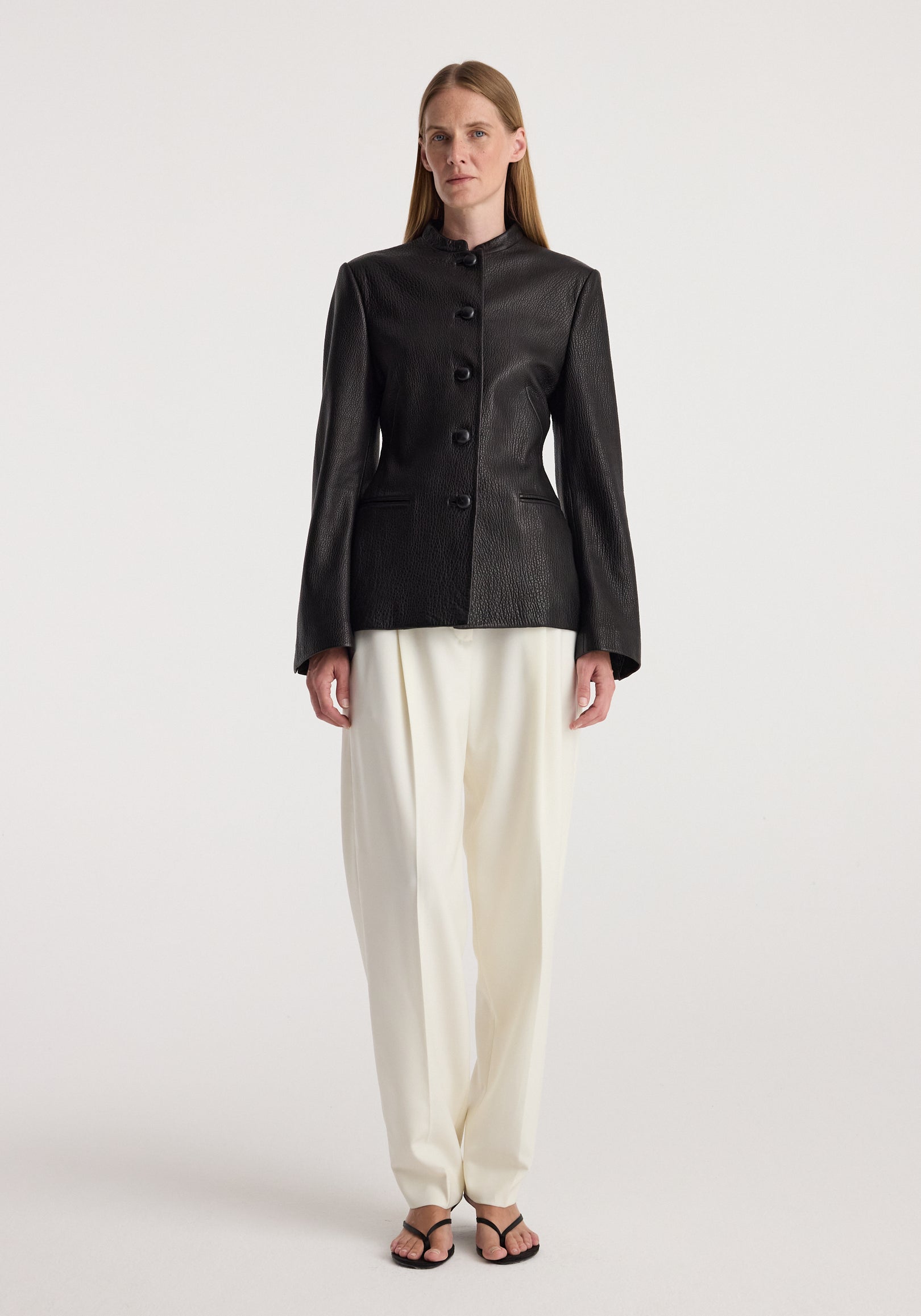 Woman wearing a tailored black shaped grain leather jacket | black with button closure, paired with loose white trousers and black sandals, standing against a plain background.