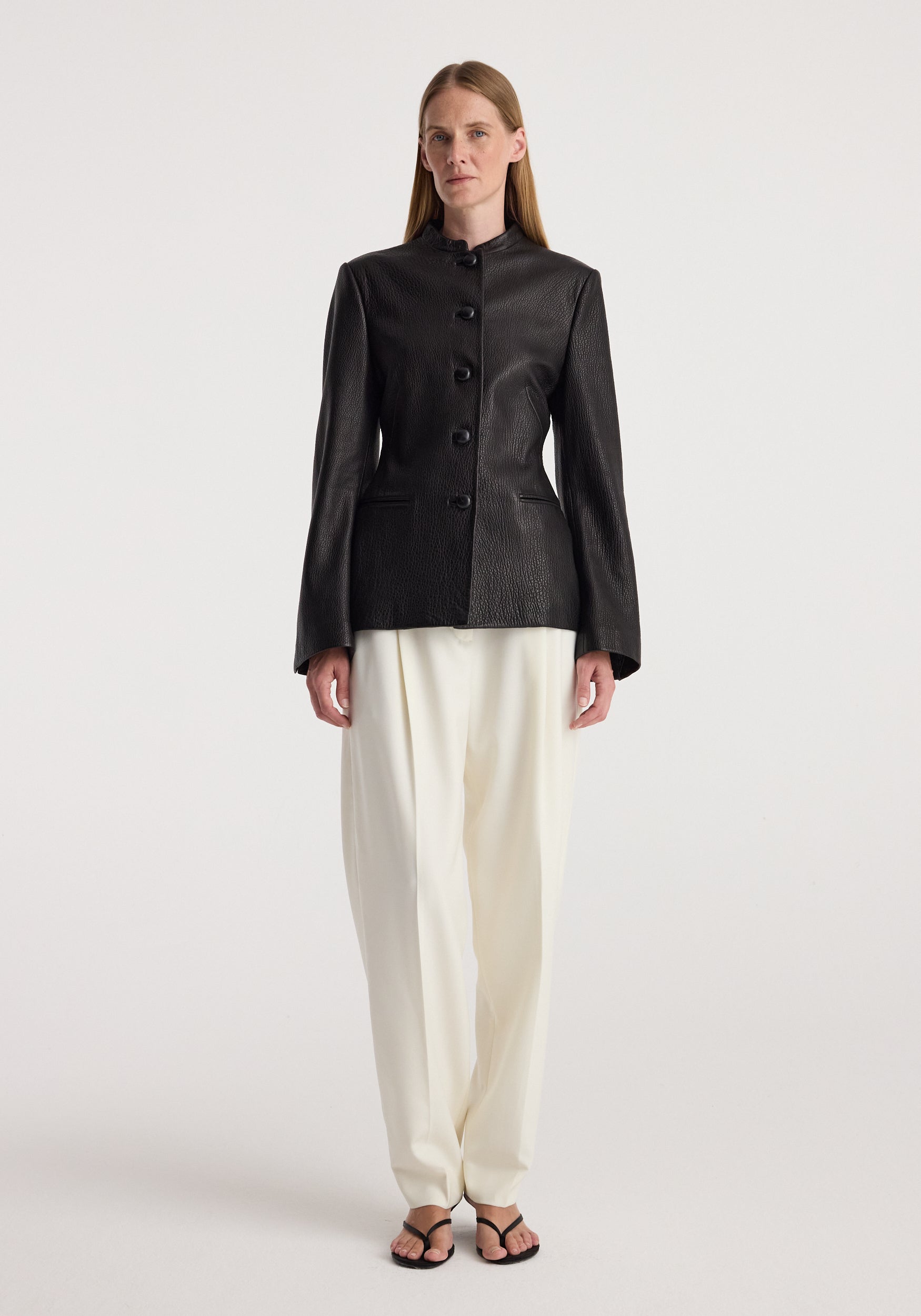 Woman wearing a tailored black shaped grain leather jacket | black with button closure, paired with loose white trousers and black sandals, standing against a plain background.