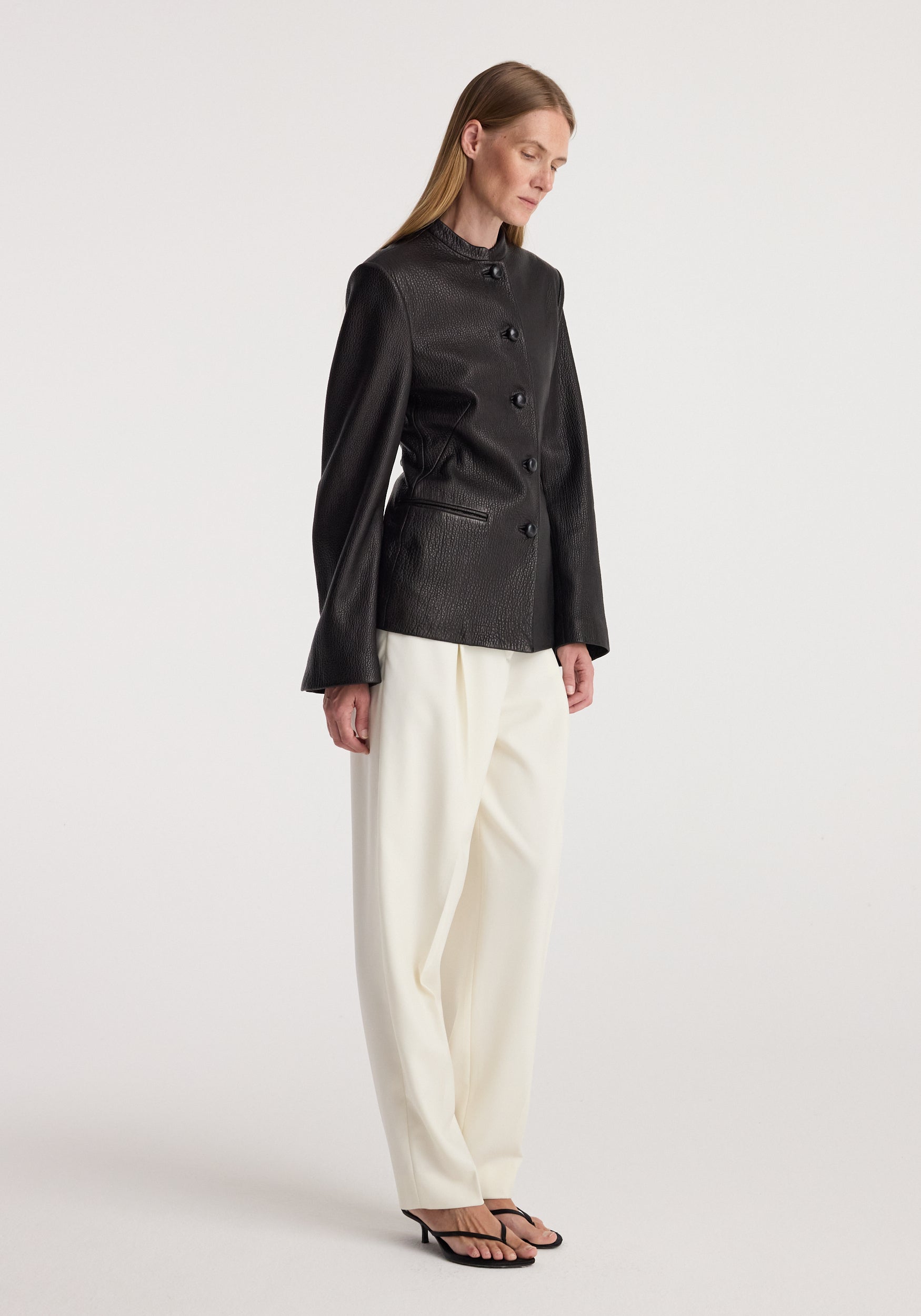 Woman wearing a fitted black shaped grain leather jacket | black with button closure, paired with loose white trousers and black open-toe heels, standing against a plain background.