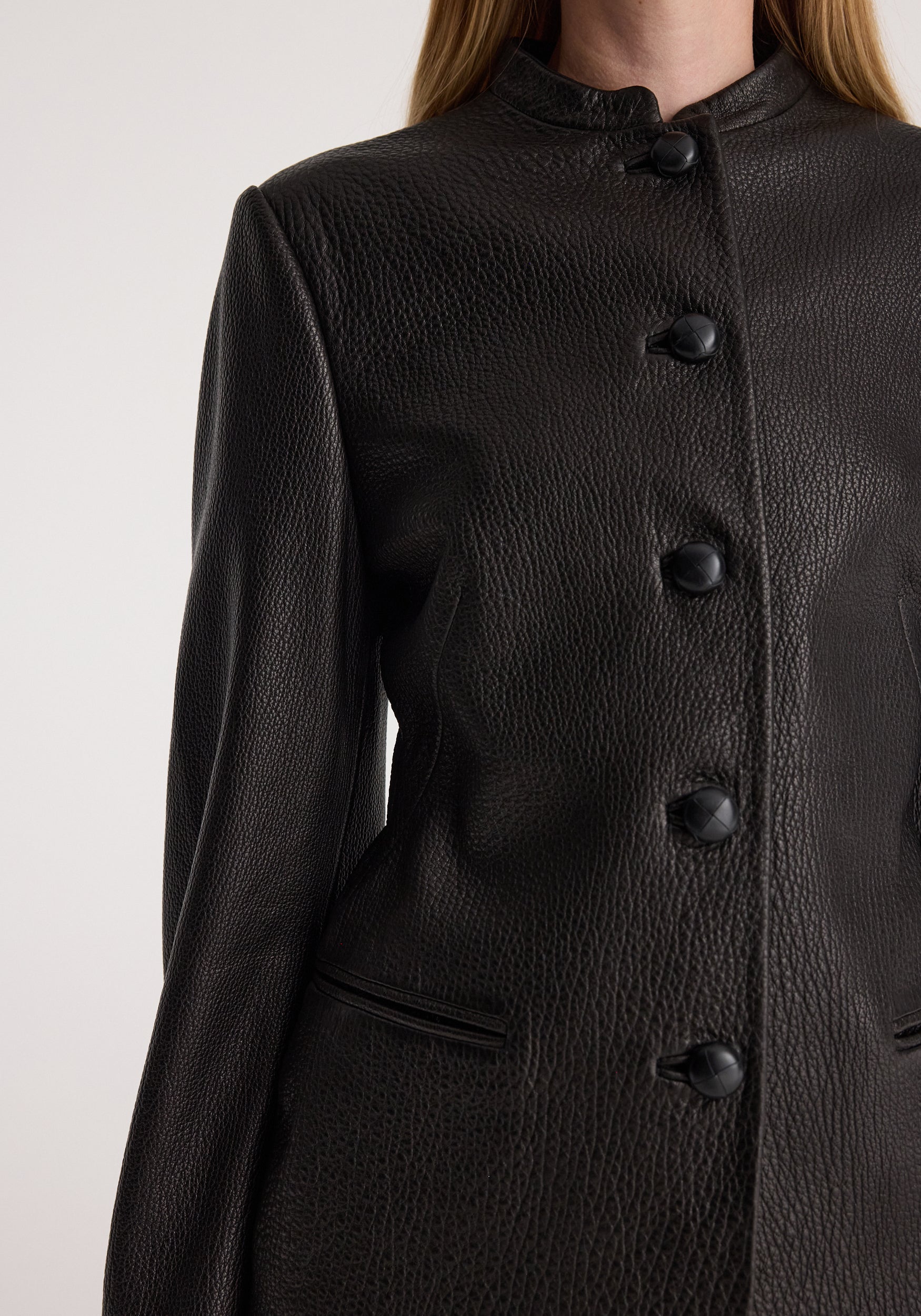 Close-up of a person wearing a black shaped grain leather jacket | black featuring textured leather, distinctive buttons, and a high collar.