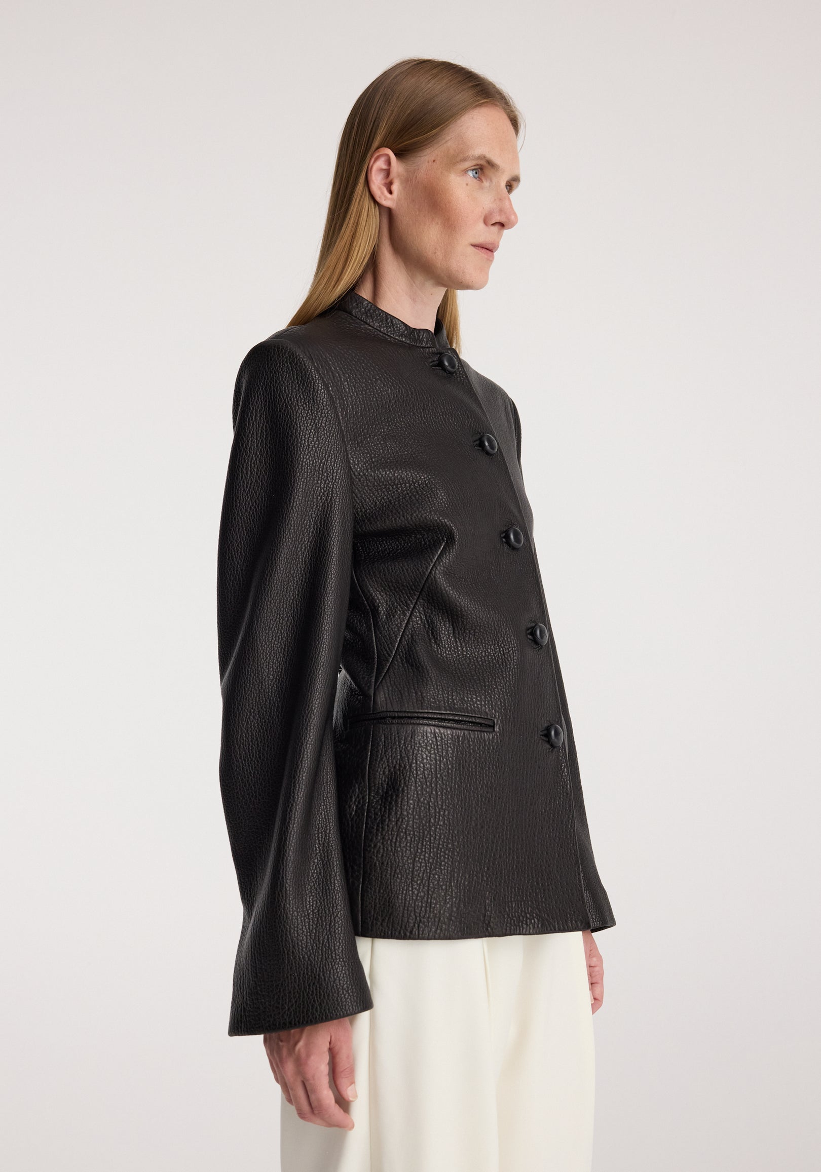 Woman wearing a black Shaped grain leather jacket | black with a high collar and button closure, paired with light-colored pants against a plain background.
