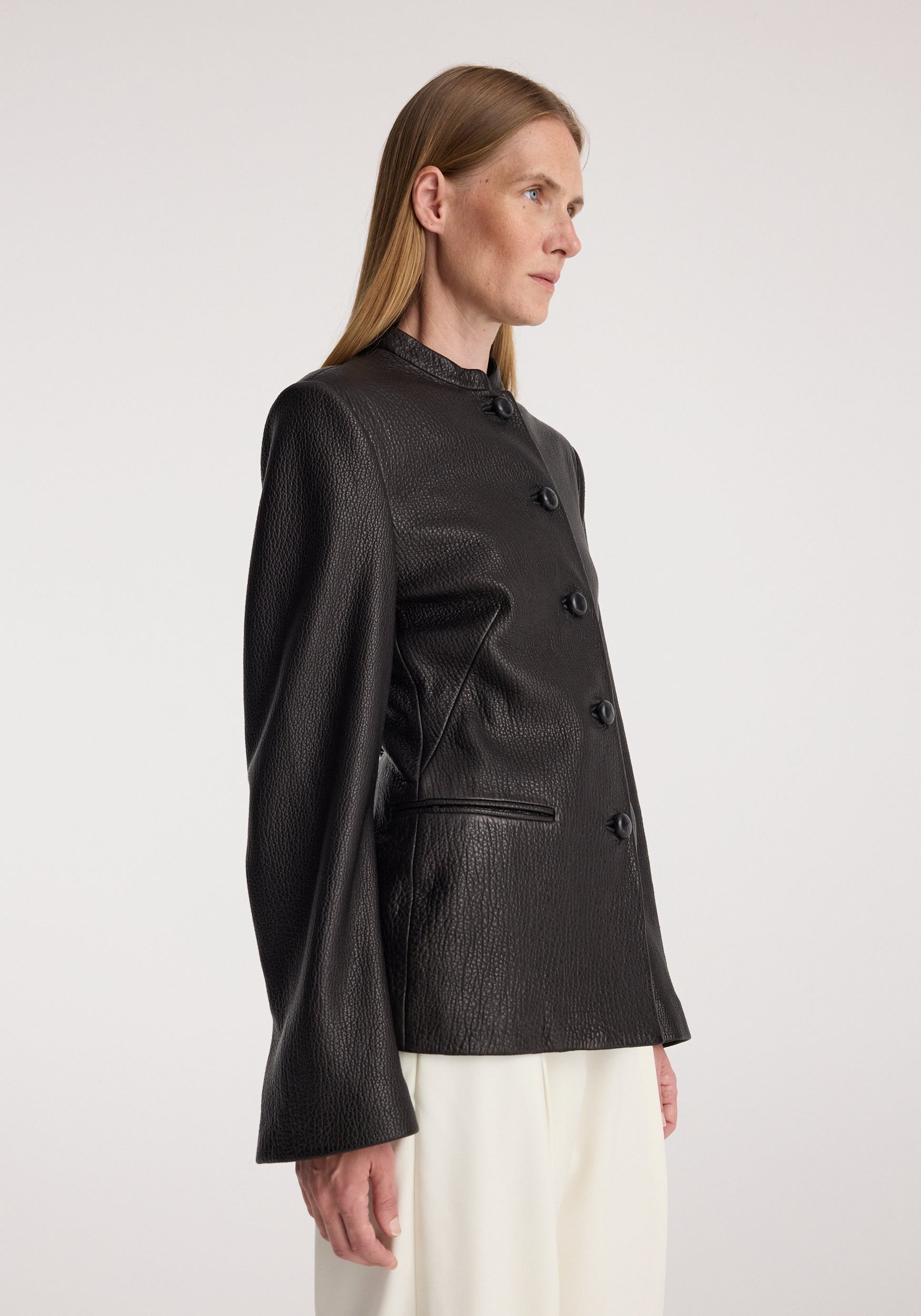 Woman wearing a black Shaped grain leather jacket | black with a high collar and button closure, paired with light-colored pants against a plain background.