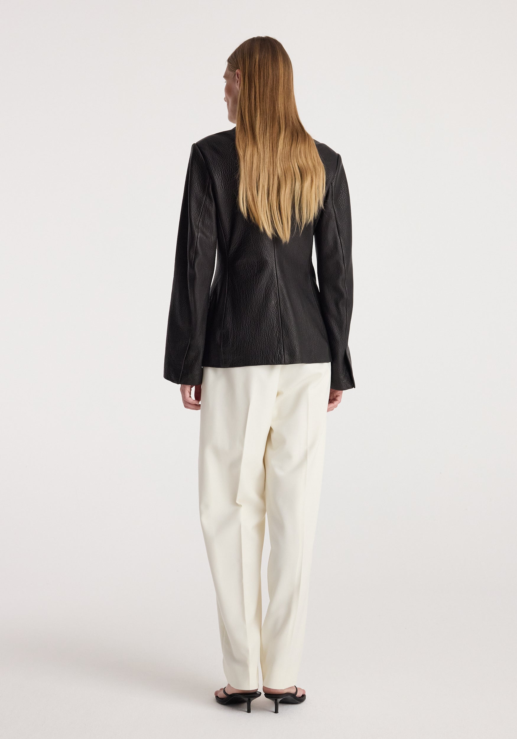 Woman wearing a shaped grain leather jacket | black, shown from the back, paired with white wide-leg trousers and black heeled sandals against a plain background.