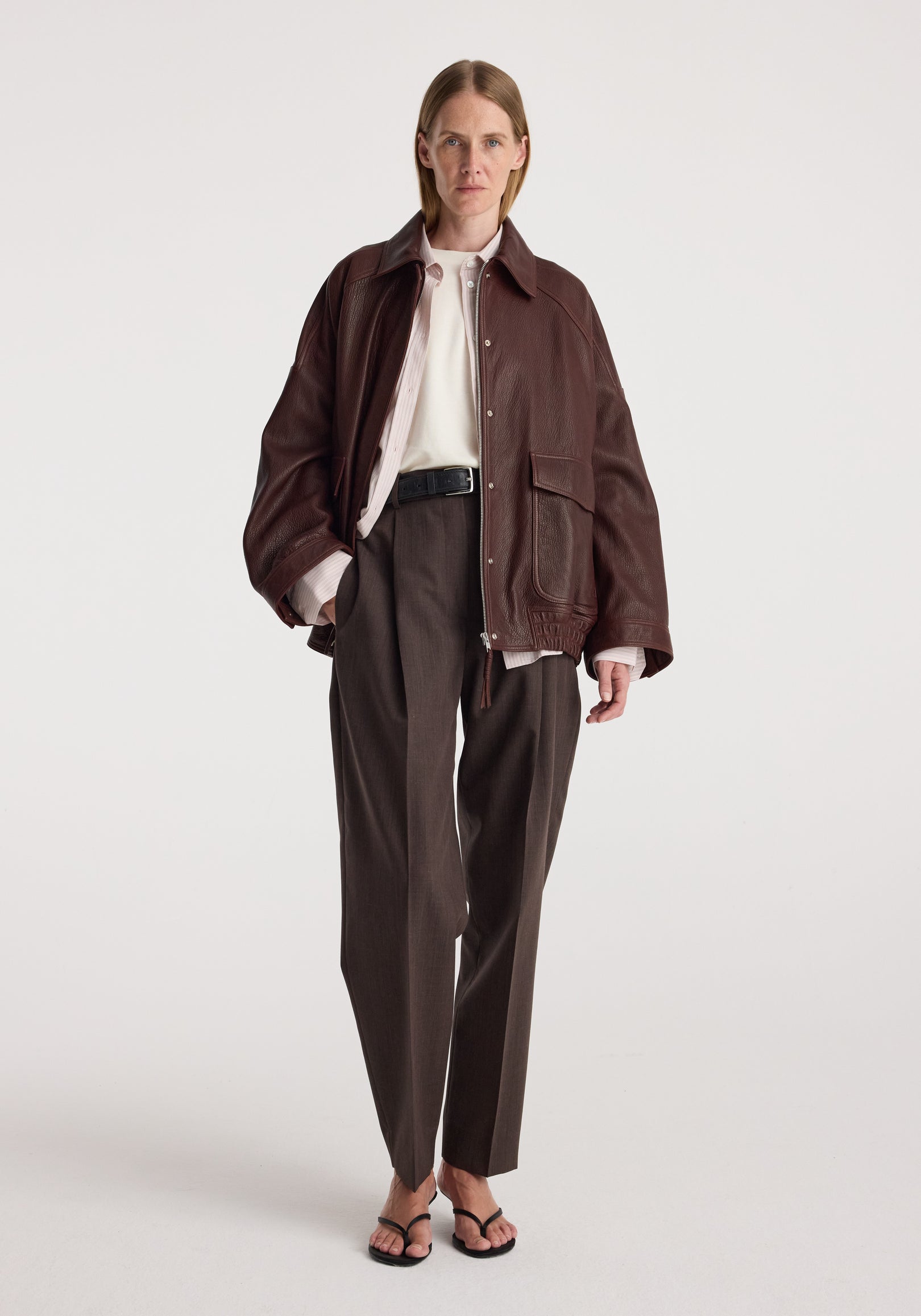 Model wearing Grain leather aviator jacket | chestnut over a white shirt with dark trousers and strappy sandals against a neutral studio background