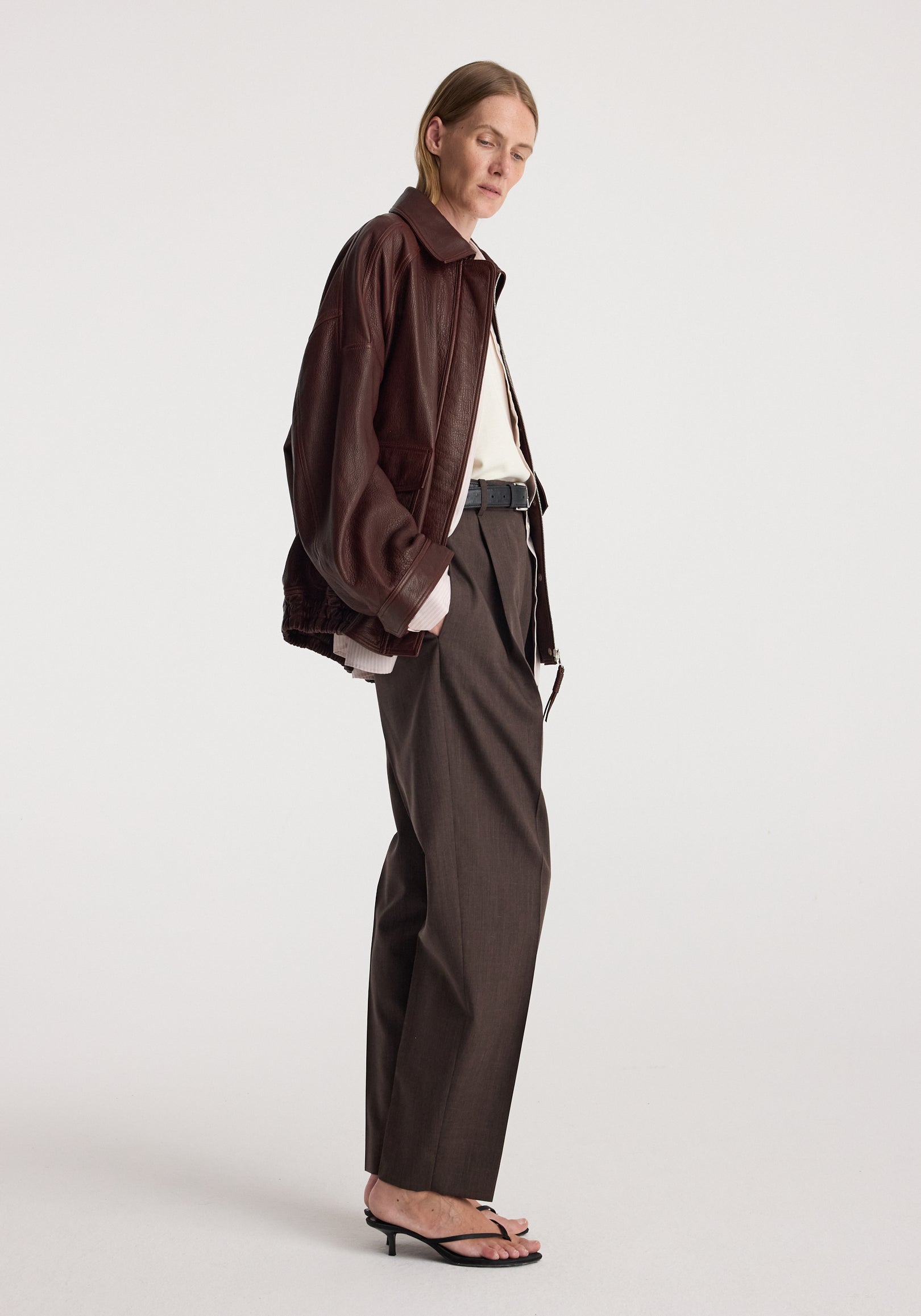 Grain leather aviator jacket | chestnut worn by a model in side profile with tailored trousers and strappy sandals against a light studio background