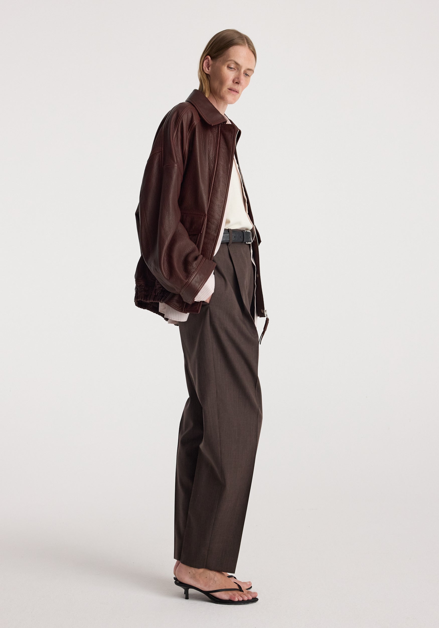 Grain leather aviator jacket | chestnut worn by a model in side profile with tailored trousers and strappy sandals against a light studio background