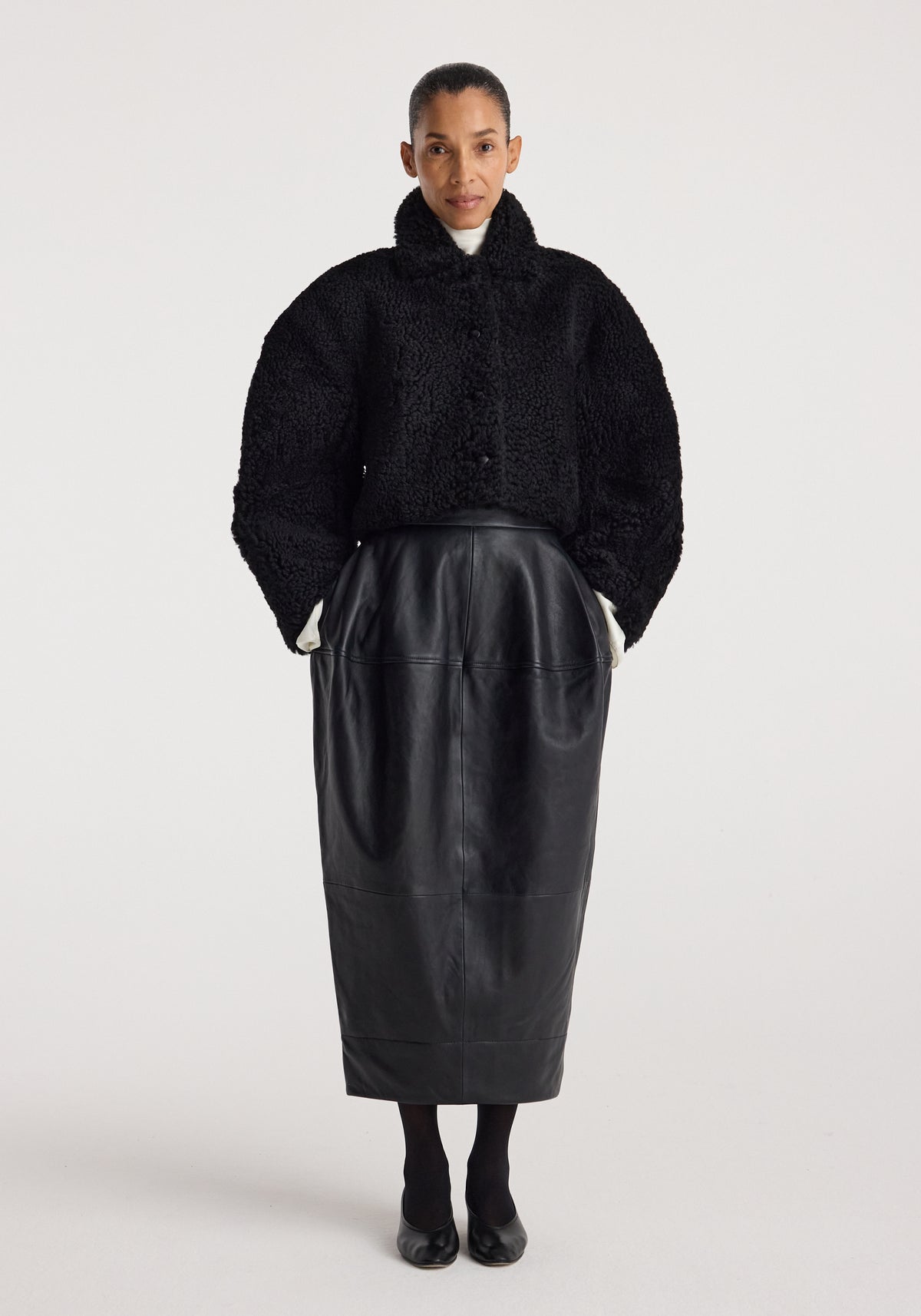 Woman wearing a black rounded shearling jacket | black with a high collar, paired with a black midi leather skirt and black shoes against a plain background.