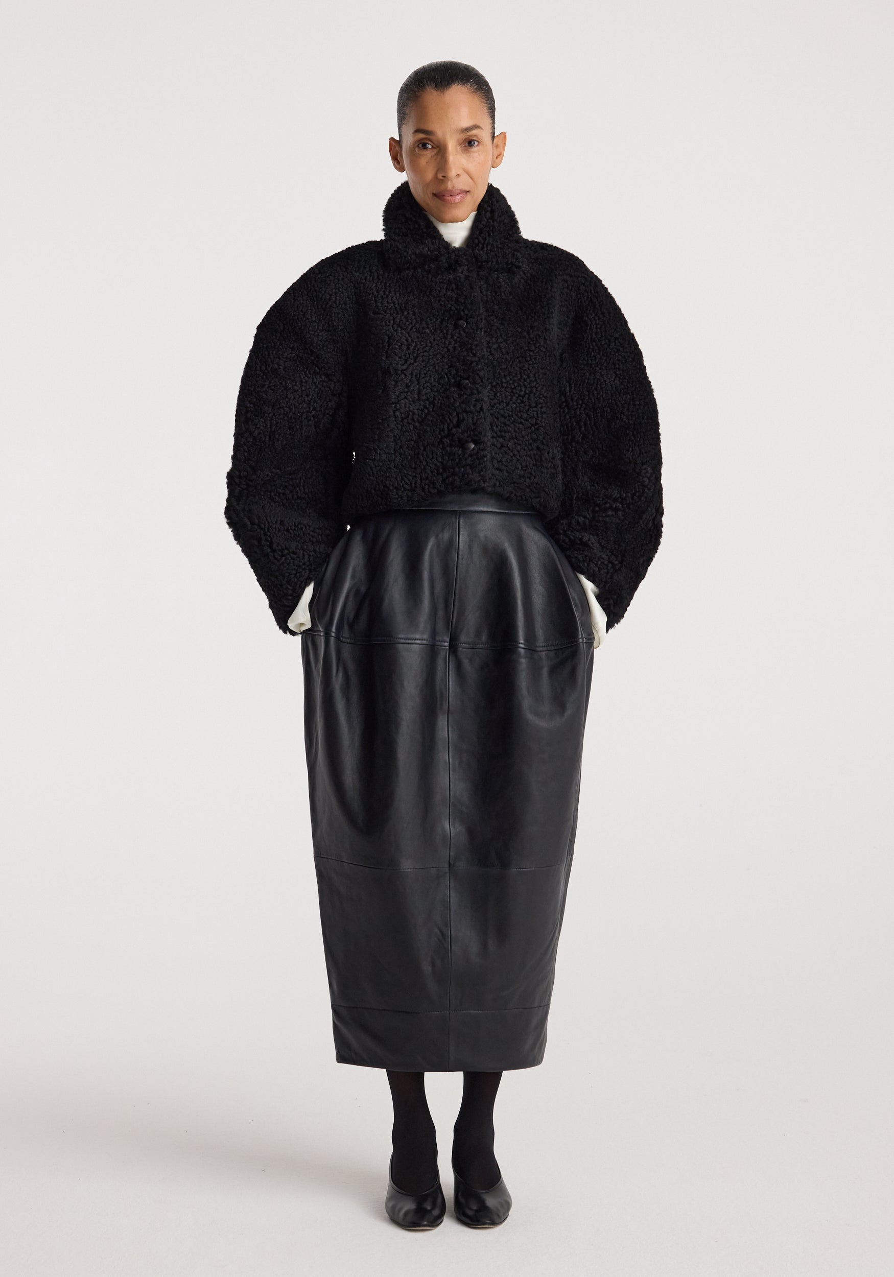 Woman wearing a black rounded shearling jacket | black with a high collar, paired with a black midi leather skirt and black shoes against a plain background.