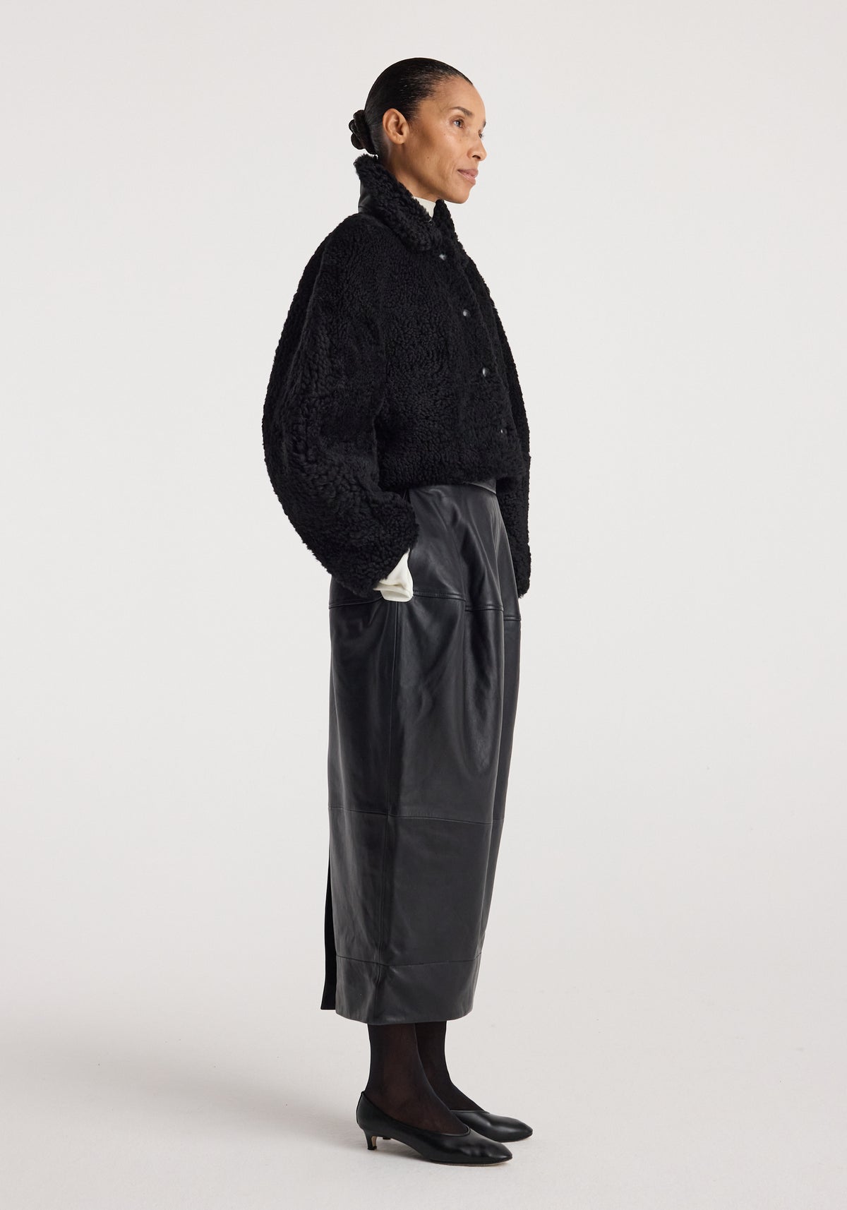 Side profile of a woman wearing a black rounded shearling jacket | black with a high collar, paired with a black leather midi skirt, black tights, and black low-heeled shoes.