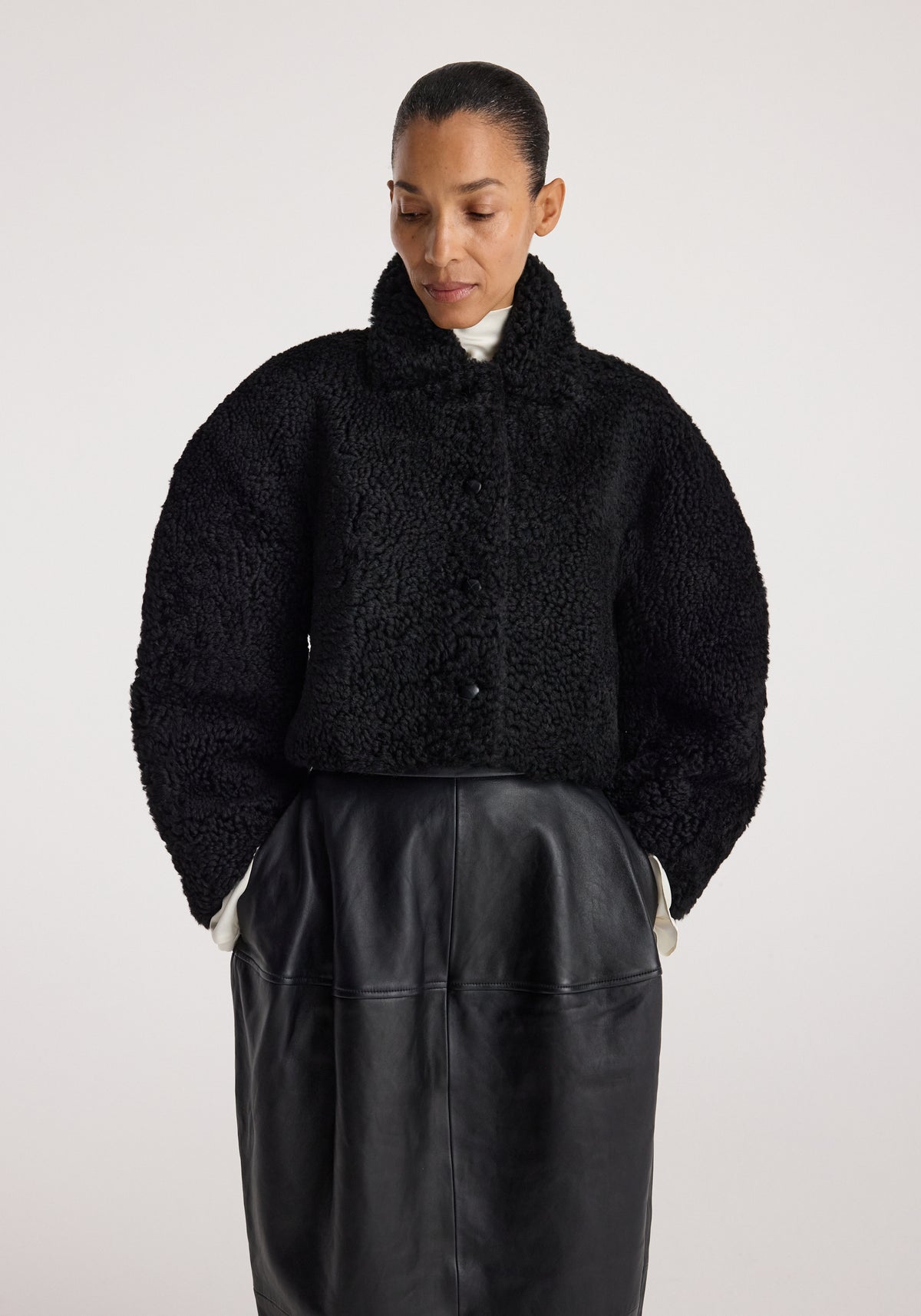 Woman wearing a black rounded shearling jacket | black with a high collar and button closure, paired with a high-waisted black leather skirt, standing against a plain background.