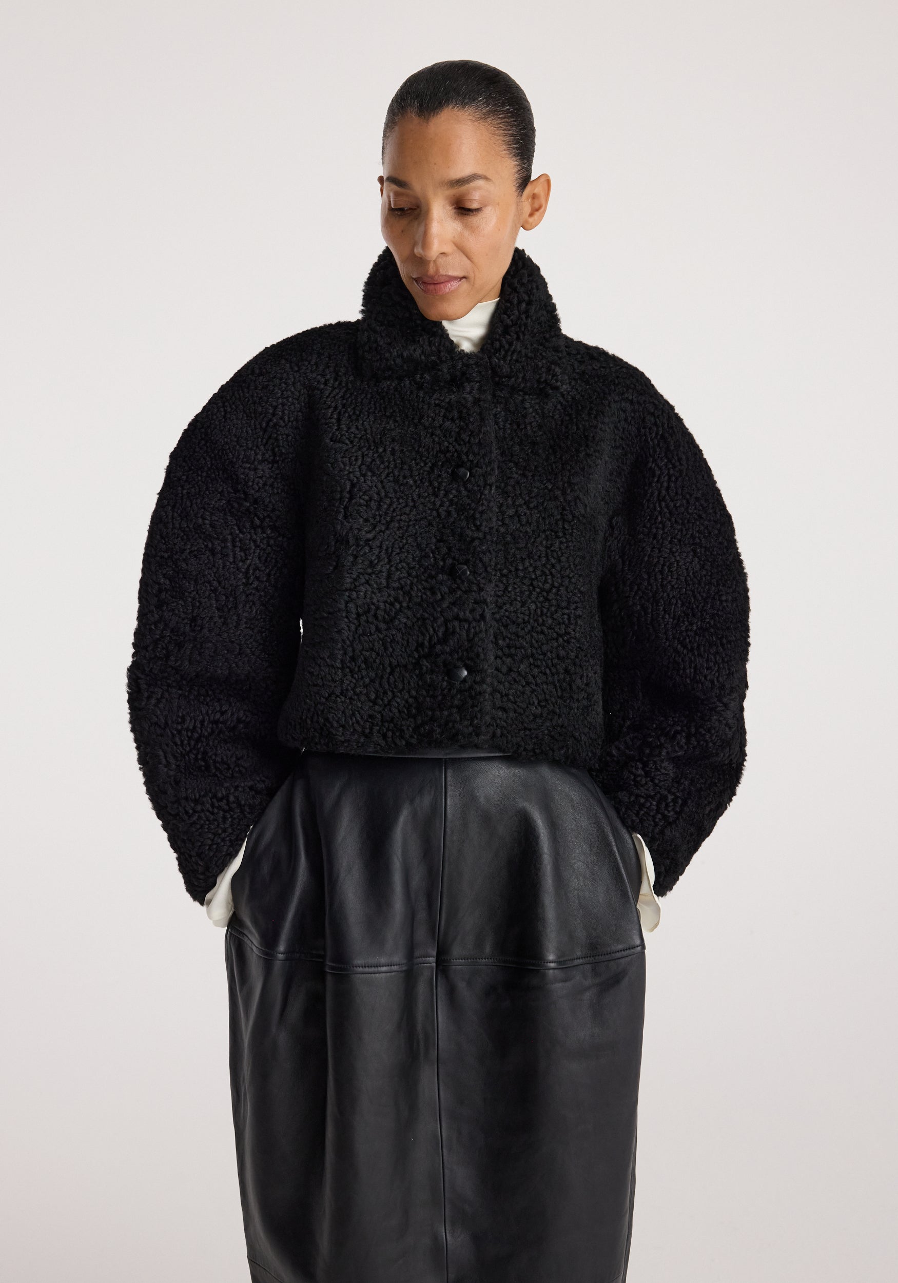 Woman wearing a black rounded shearling jacket | black with a high collar and button closure, paired with a high-waisted black leather skirt, standing against a plain background.