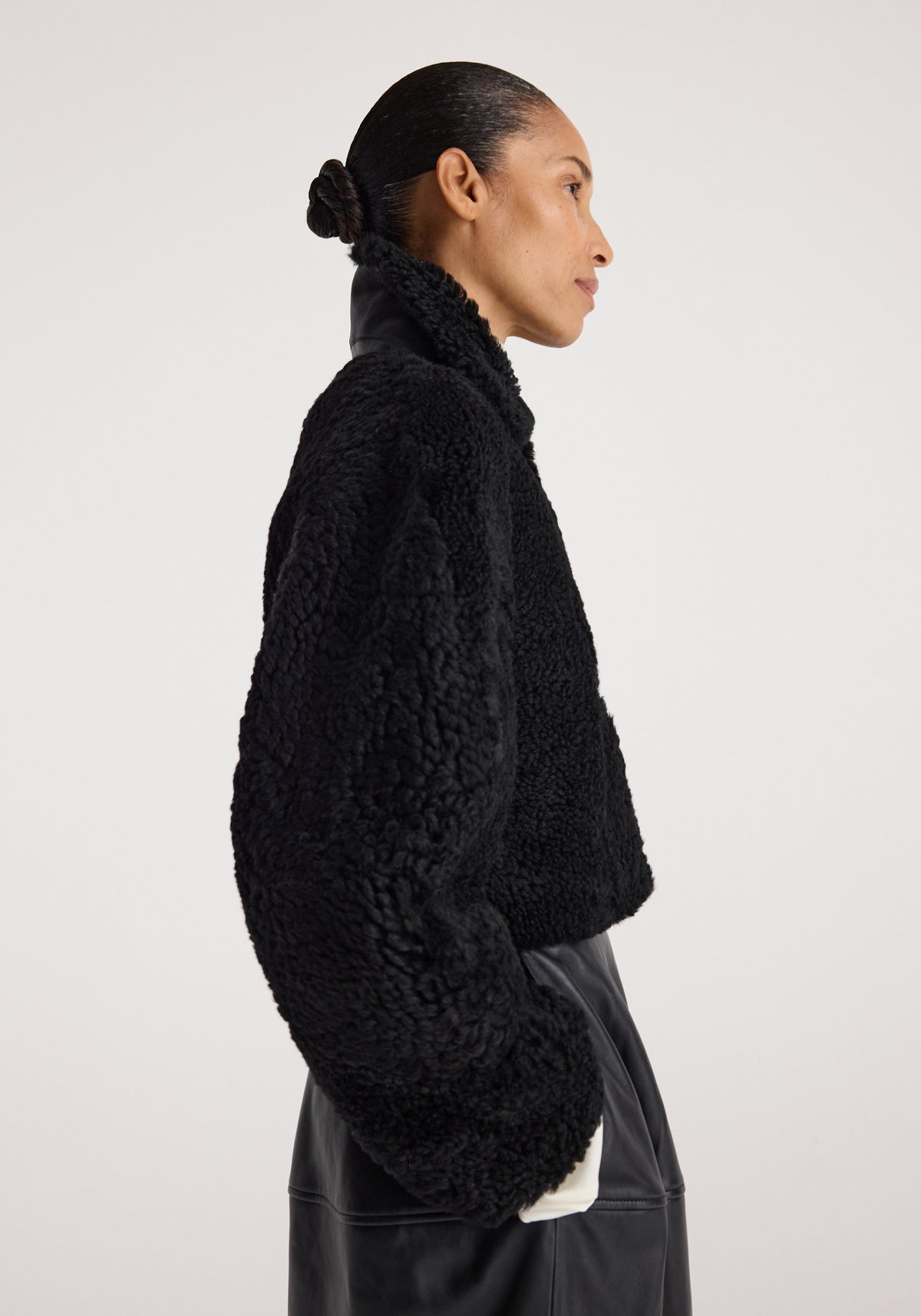 Side profile of a person wearing a black rounded shearling jacket | black with a textured, cozy finish and a high collar against a plain background.