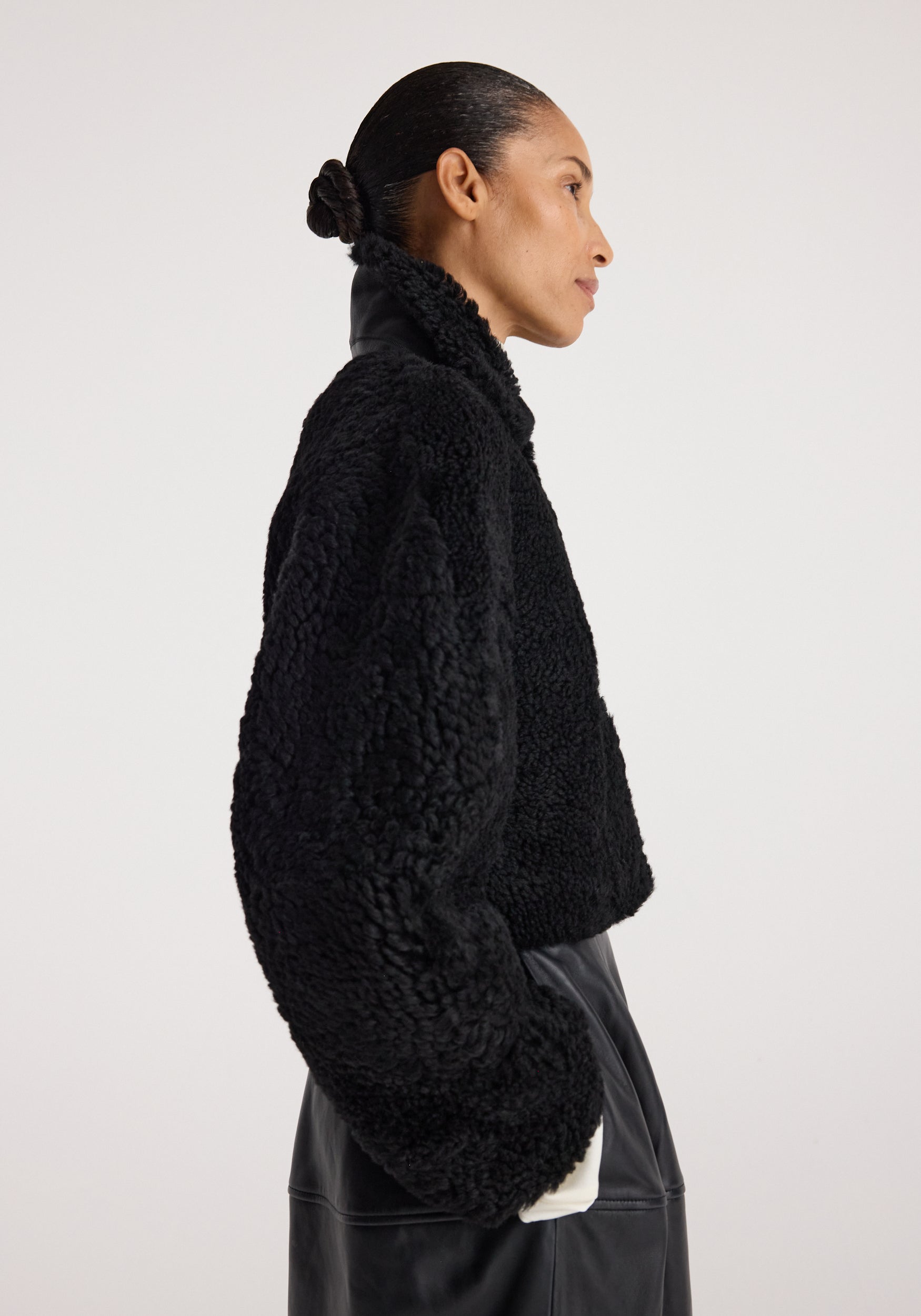 Side profile of a person wearing a black rounded shearling jacket | black with a textured, cozy finish and a high collar against a plain background.