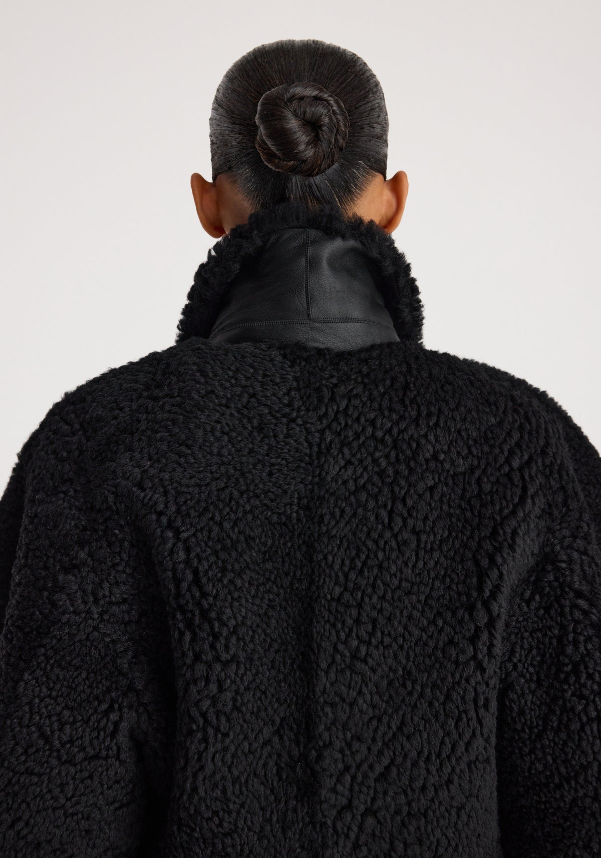 Back view of person with hair in a bun wearing a textured Rounded shearling jacket | black with a high collar against a plain background.