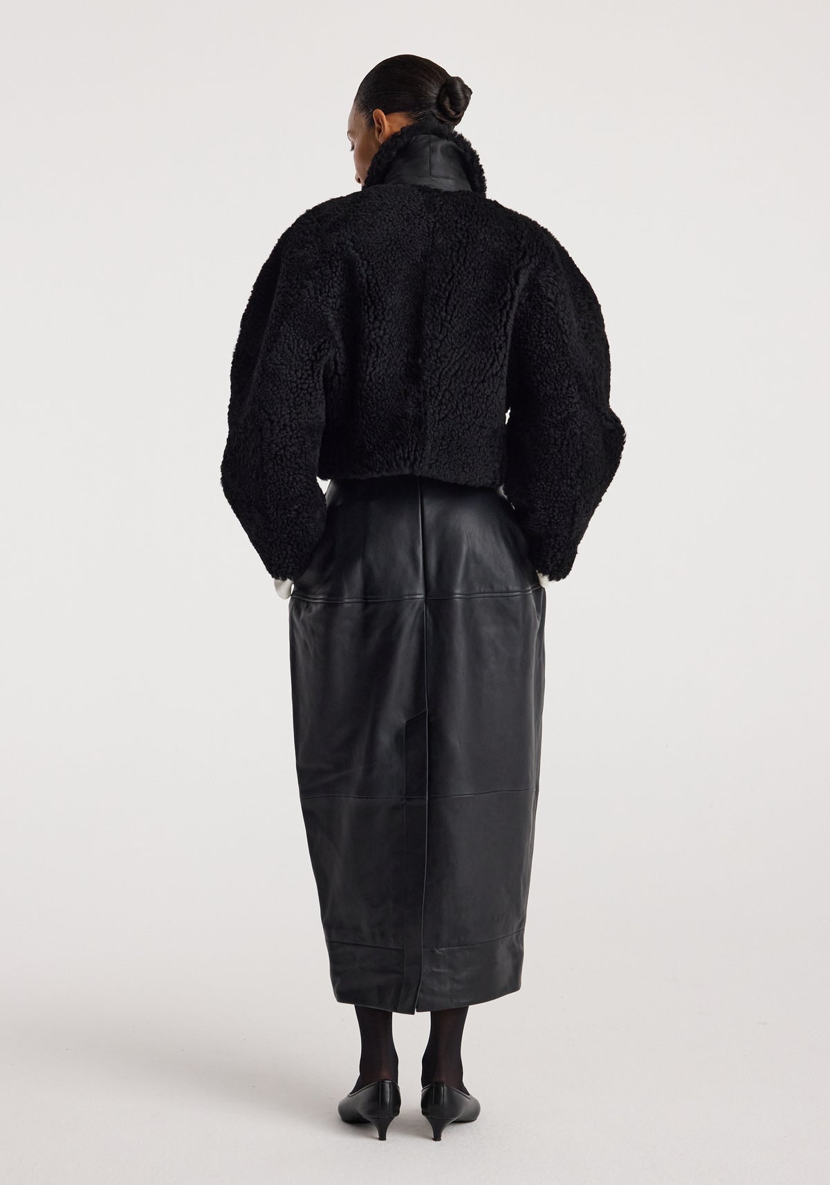 Woman wearing a black rounded shearling jacket | black with a high collar, paired with a long black leather skirt and black heels, standing against a white background.