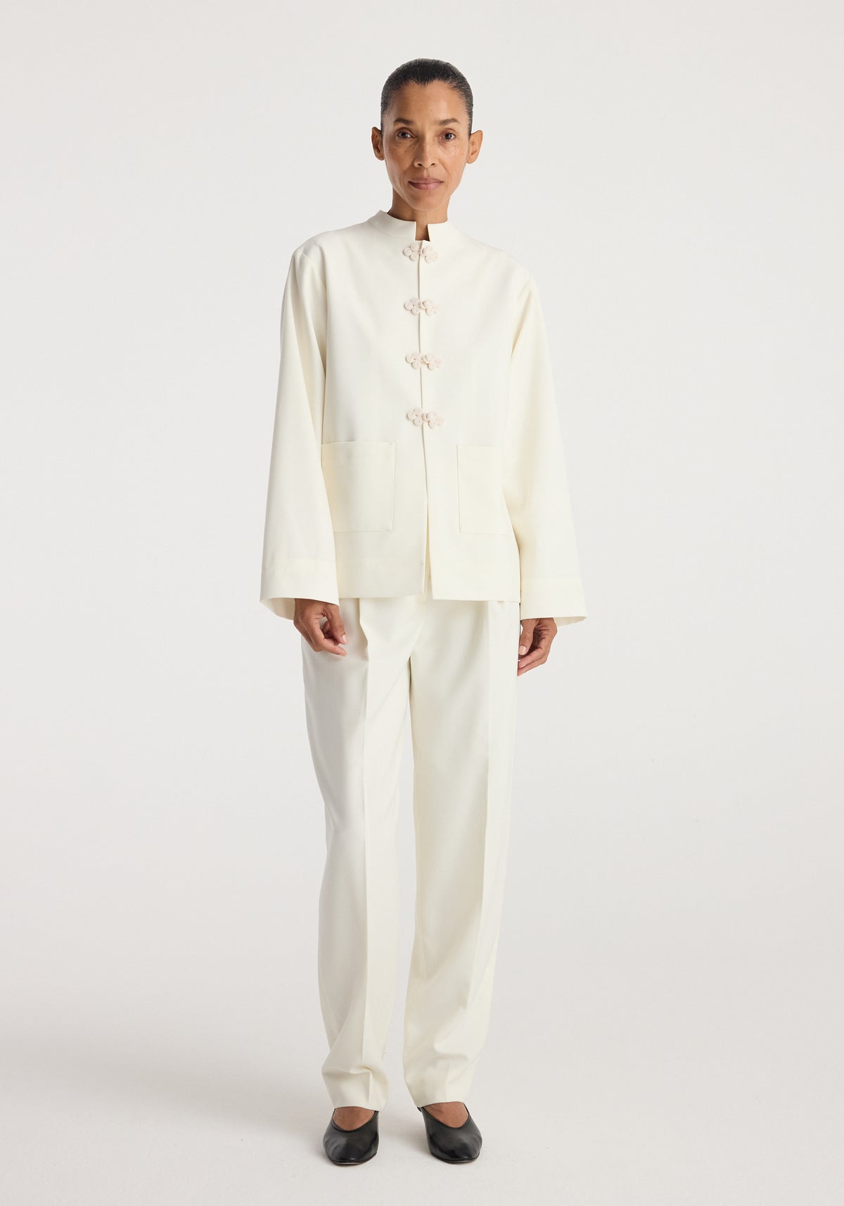 Model wearing a minimalist off-white Pankou closure jacket | off white featuring a high collar, long sleeves, front pockets, and traditional knot button closures paired with matching pants.
