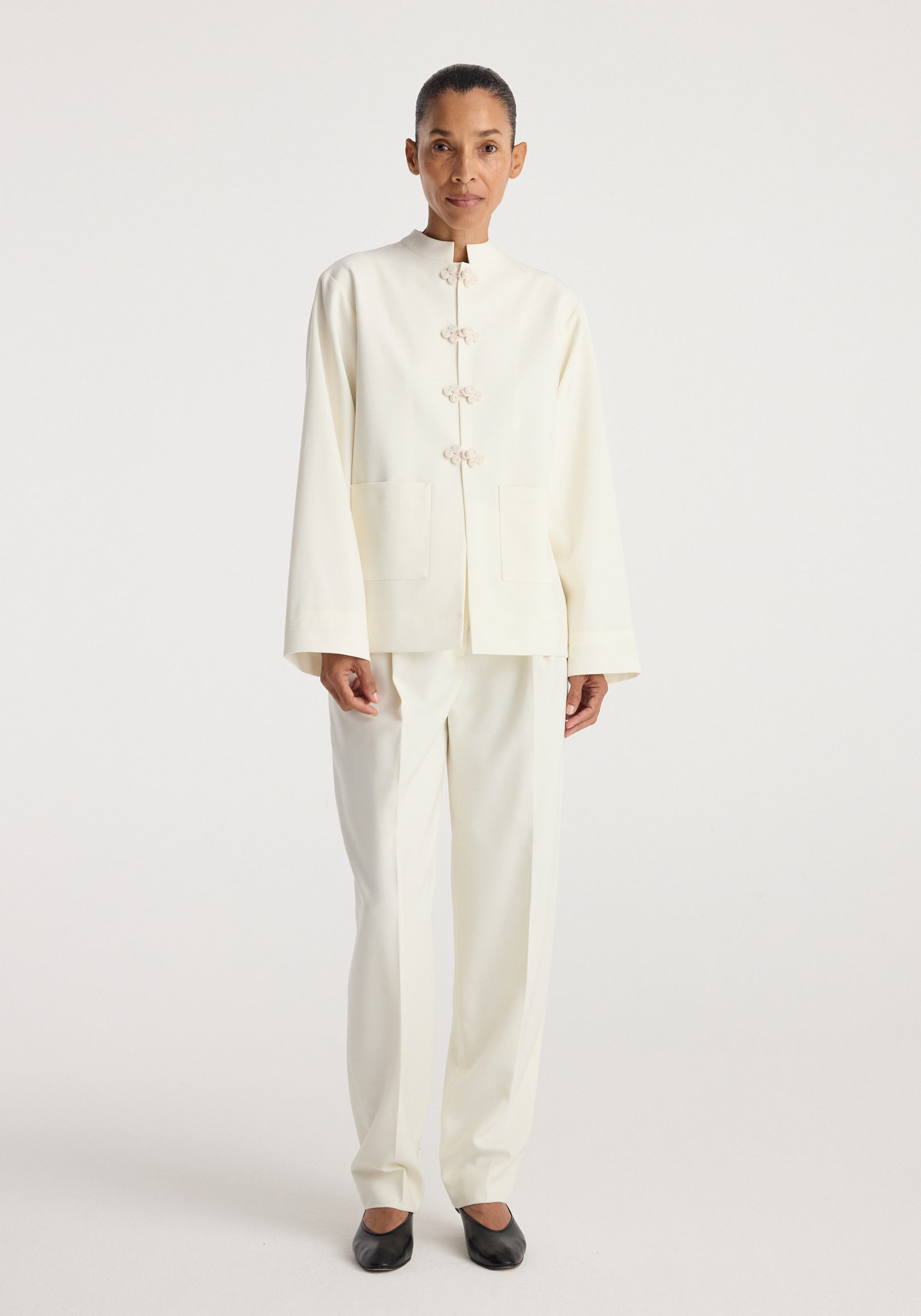 Model wearing a minimalist off-white Pankou closure jacket | off white featuring a high collar, long sleeves, front pockets, and traditional knot button closures paired with matching pants.