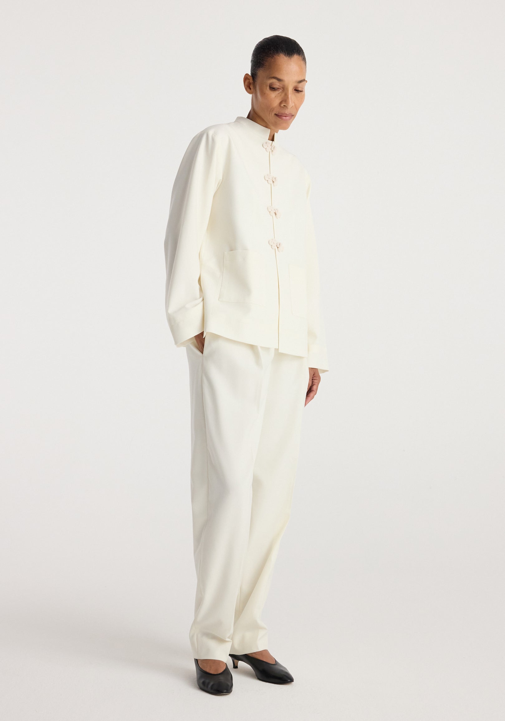 woman wearing an off white Pankou closure jacket | off white with mandarin collar and matching wide-leg pants, styled with black heels against a plain background