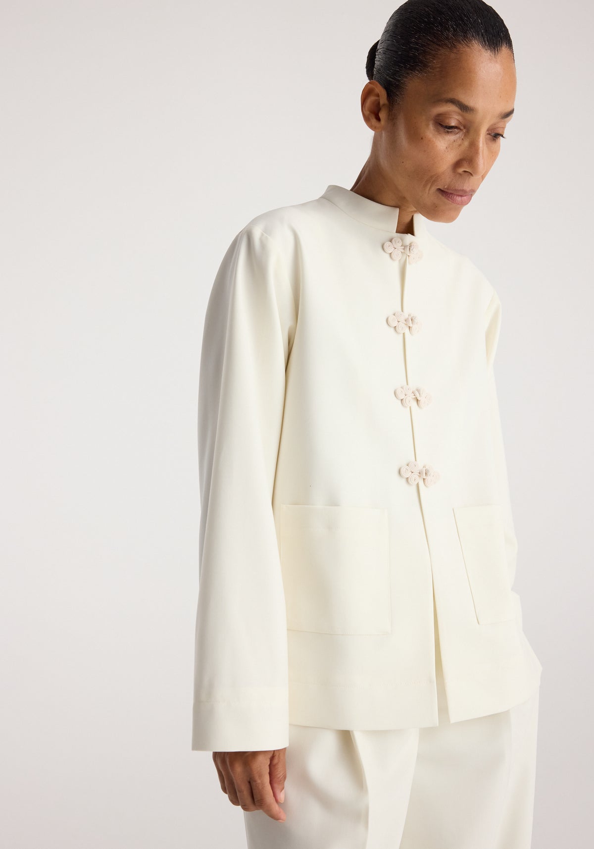 Woman wearing a minimalistic off-white Pankou closure jacket | off white with a stand collar and large front pockets, styled with matching pants against a neutral background.