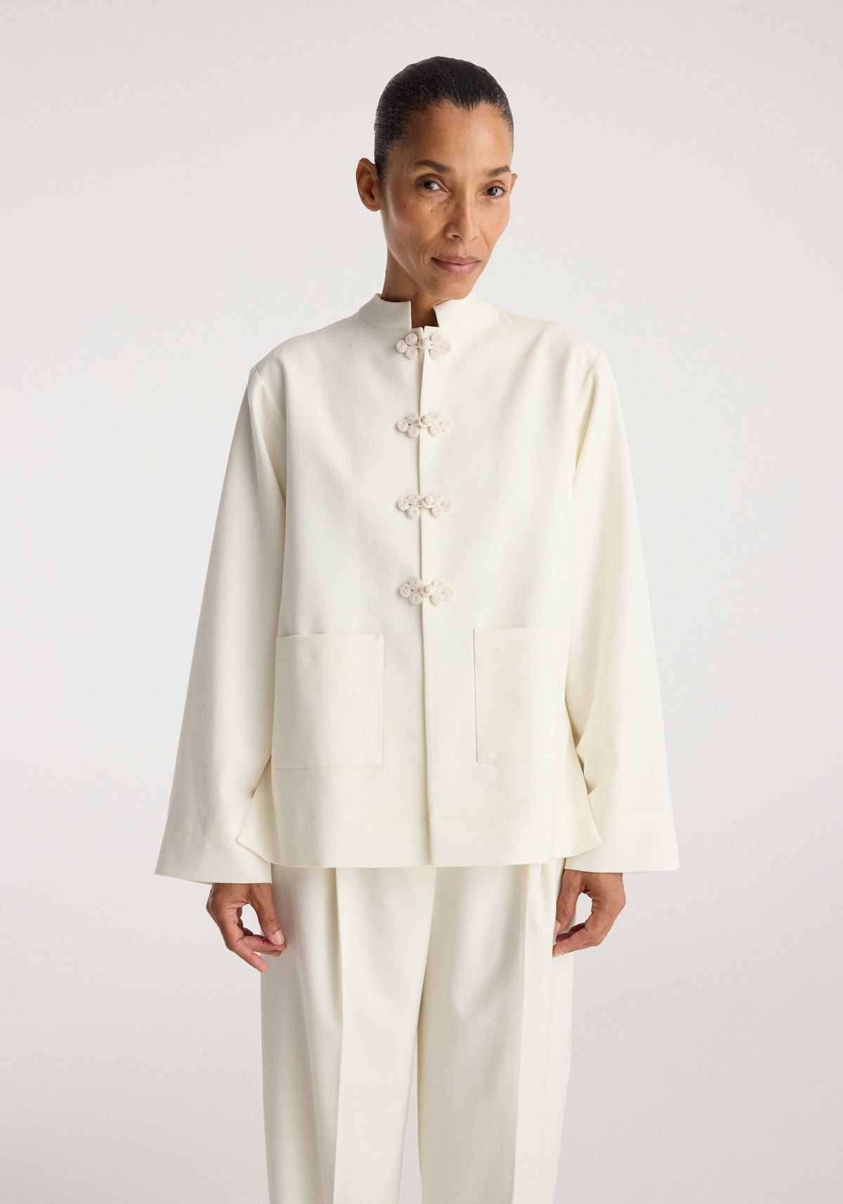 Woman wearing a minimalist off-white Pankou closure jacket | off white with traditional knot buttons and two front pockets, paired with matching off-white trousers.