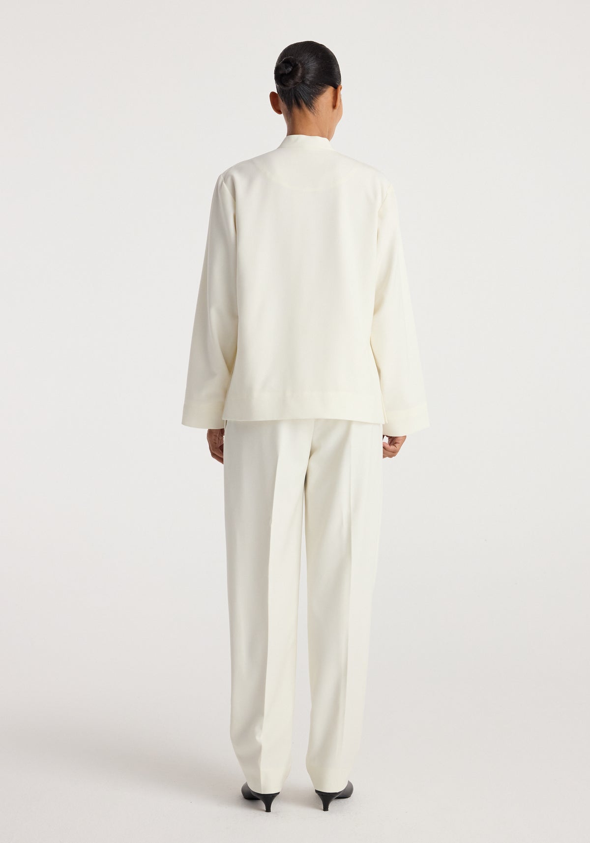 Back view of a person wearing a Pankou closure jacket | off white paired with matching off white trousers and black heeled shoes against a plain background.