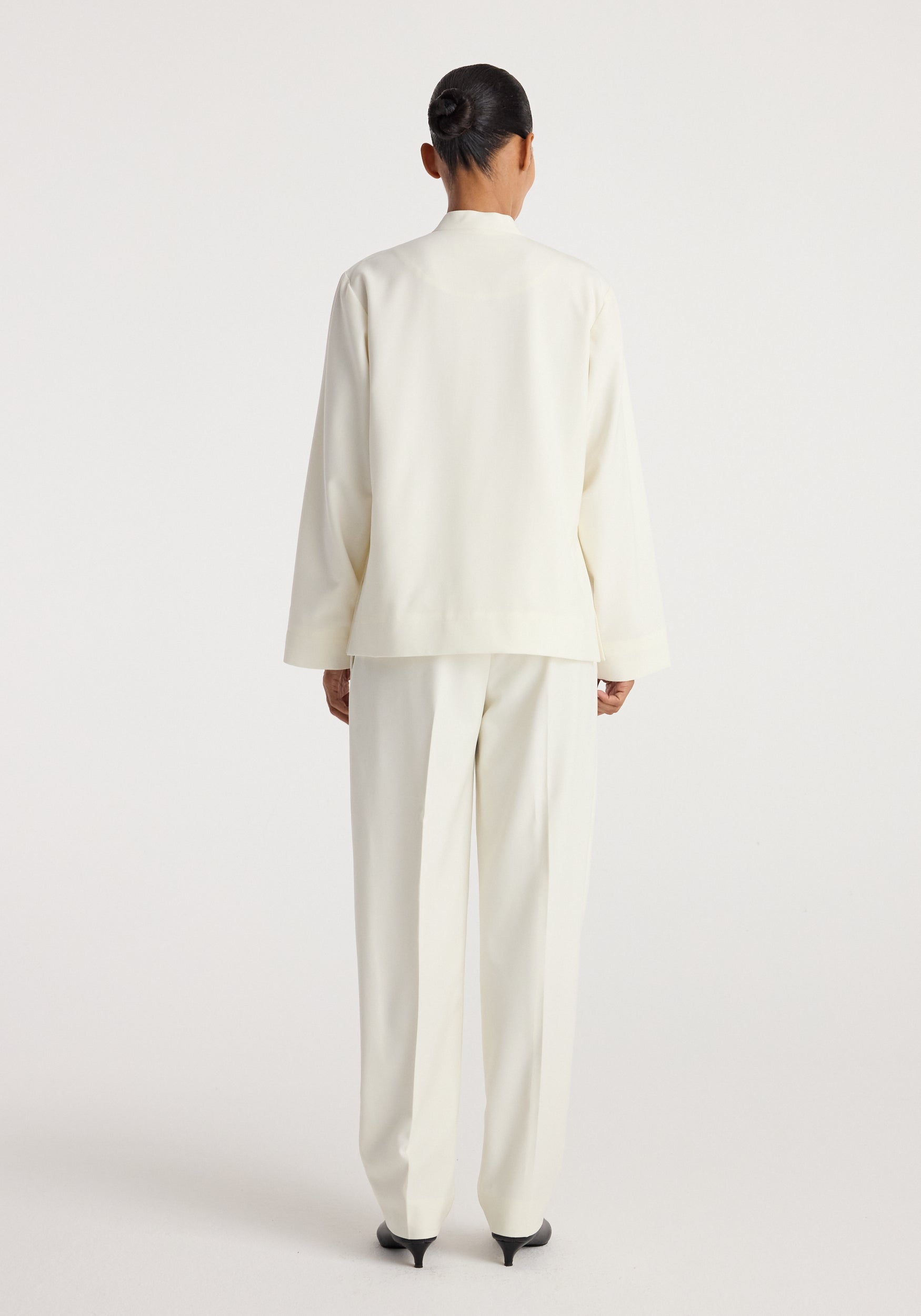 Back view of a person wearing a Pankou closure jacket | off white paired with matching off white trousers and black heeled shoes against a plain background.