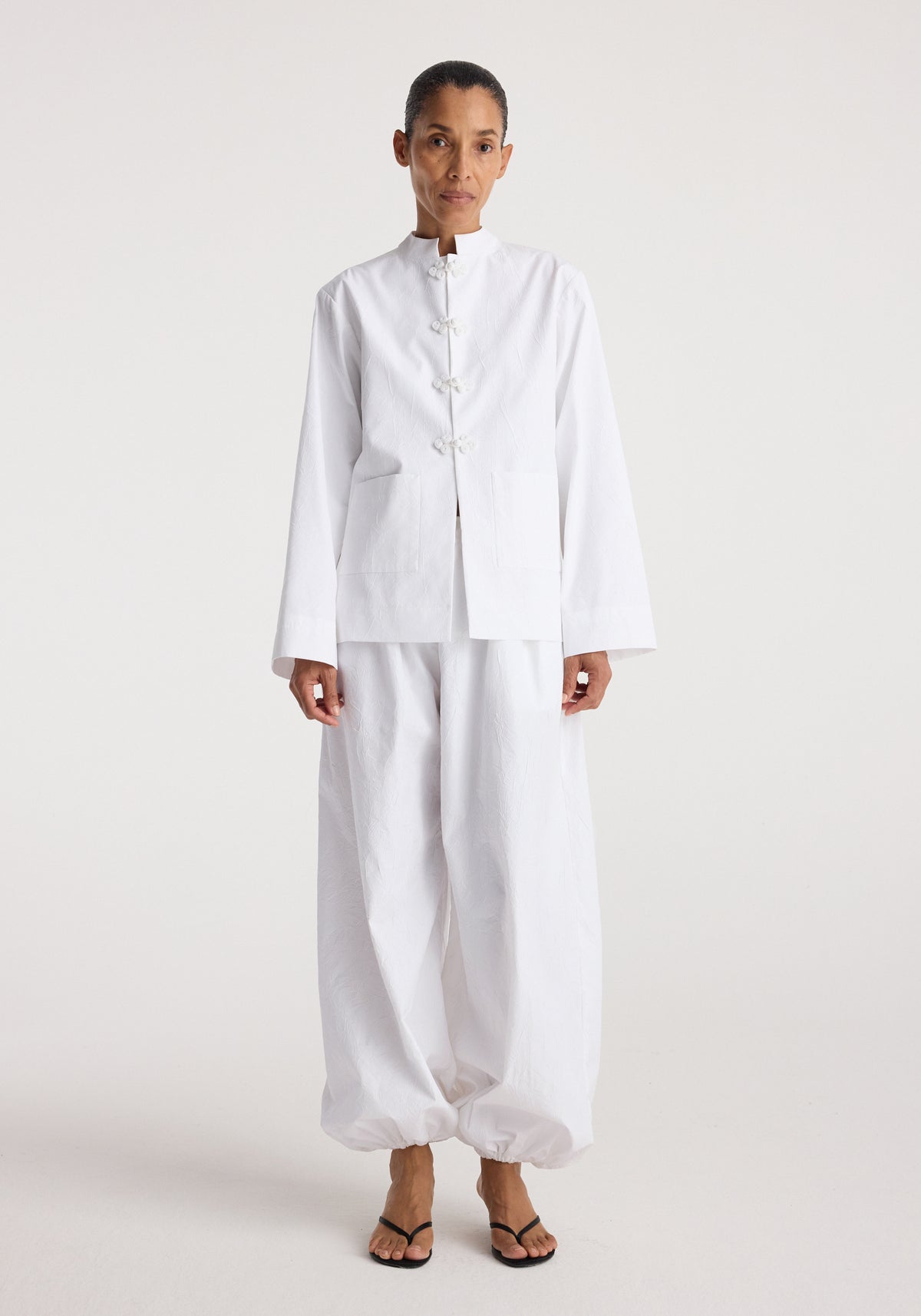 Woman wearing a minimalist white jacket with traditional pankou closures, styled with matching wide-leg pants, showcasing the Pankou closure crushed jacket | optic white.