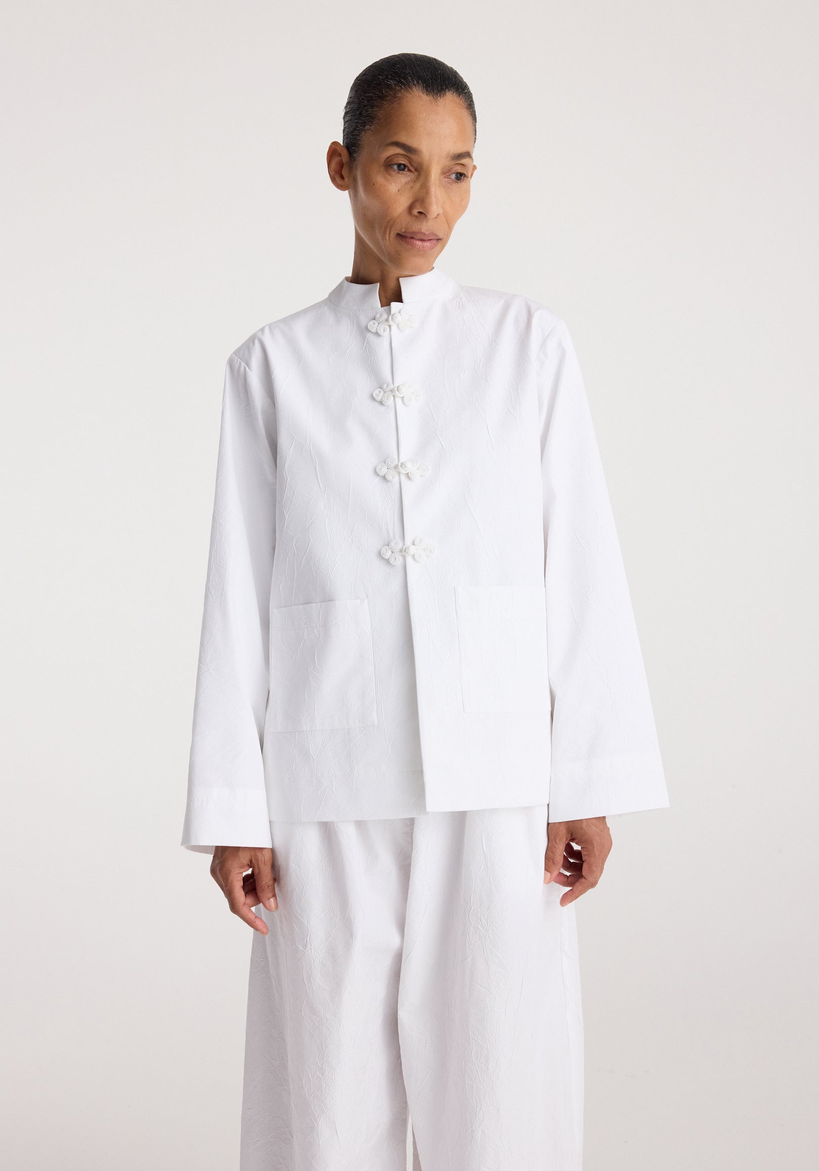 Woman wearing a clean and minimalist Pankou closure crushed jacket | optic white with traditional knot buttons and two front pockets against a plain light background.