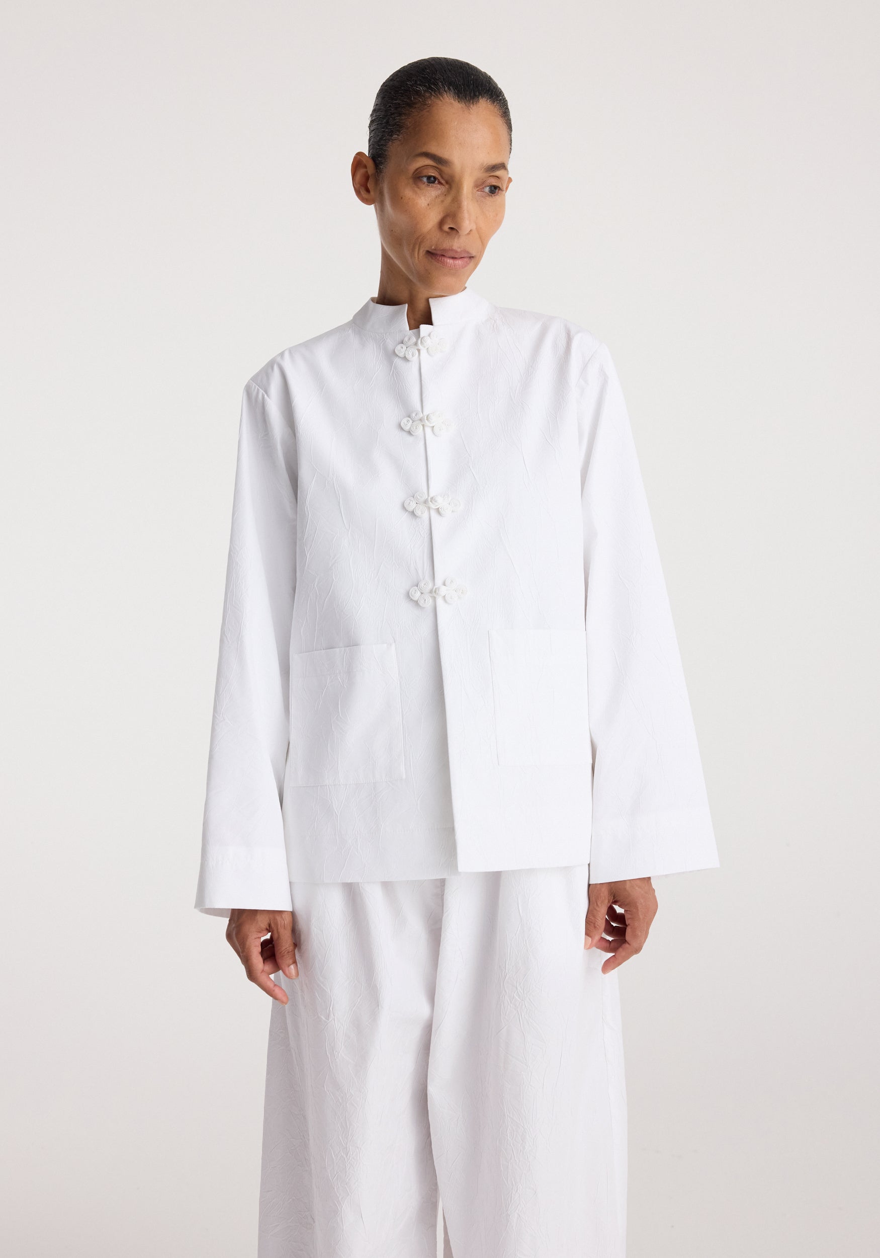 Woman wearing a clean and minimalist Pankou closure crushed jacket | optic white with traditional knot buttons and two front pockets against a plain light background.