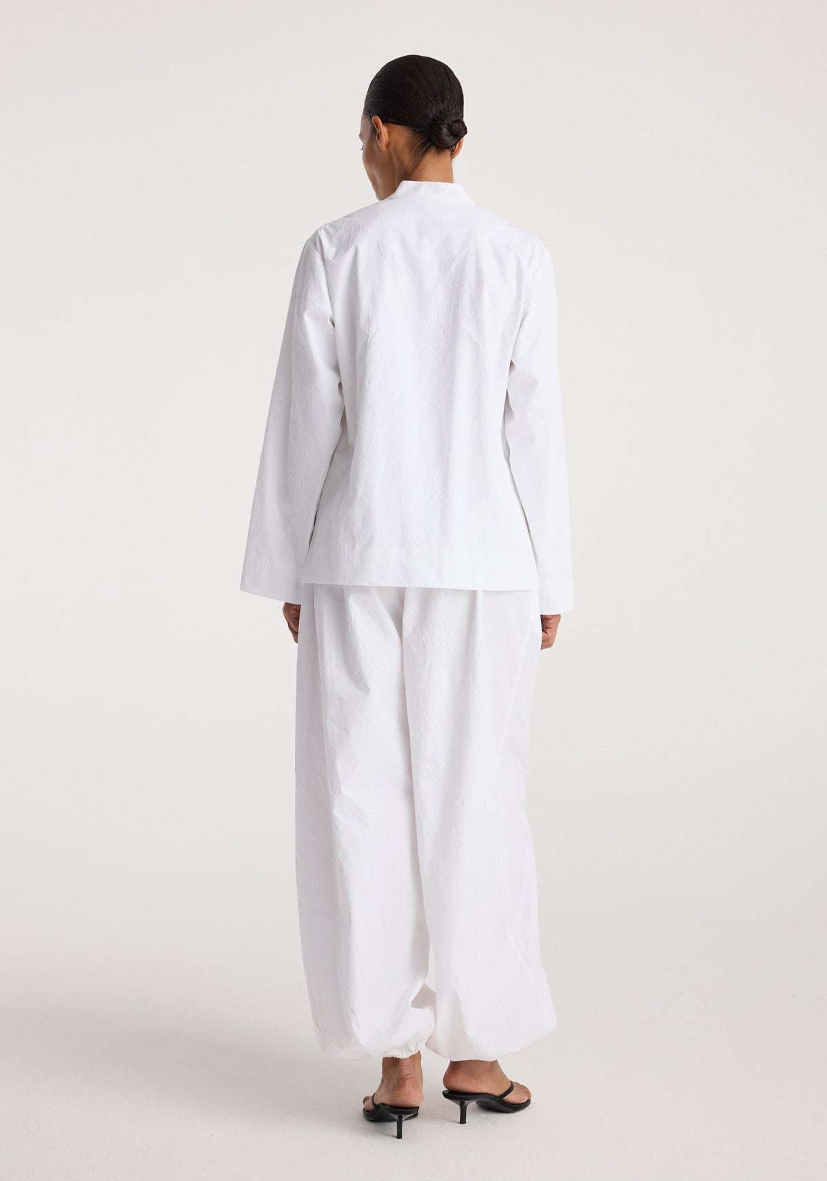 Woman wearing a Pankou closure crushed jacket | optic white, shown from the back, paired with loose white pants and black heeled sandals on a plain background.
