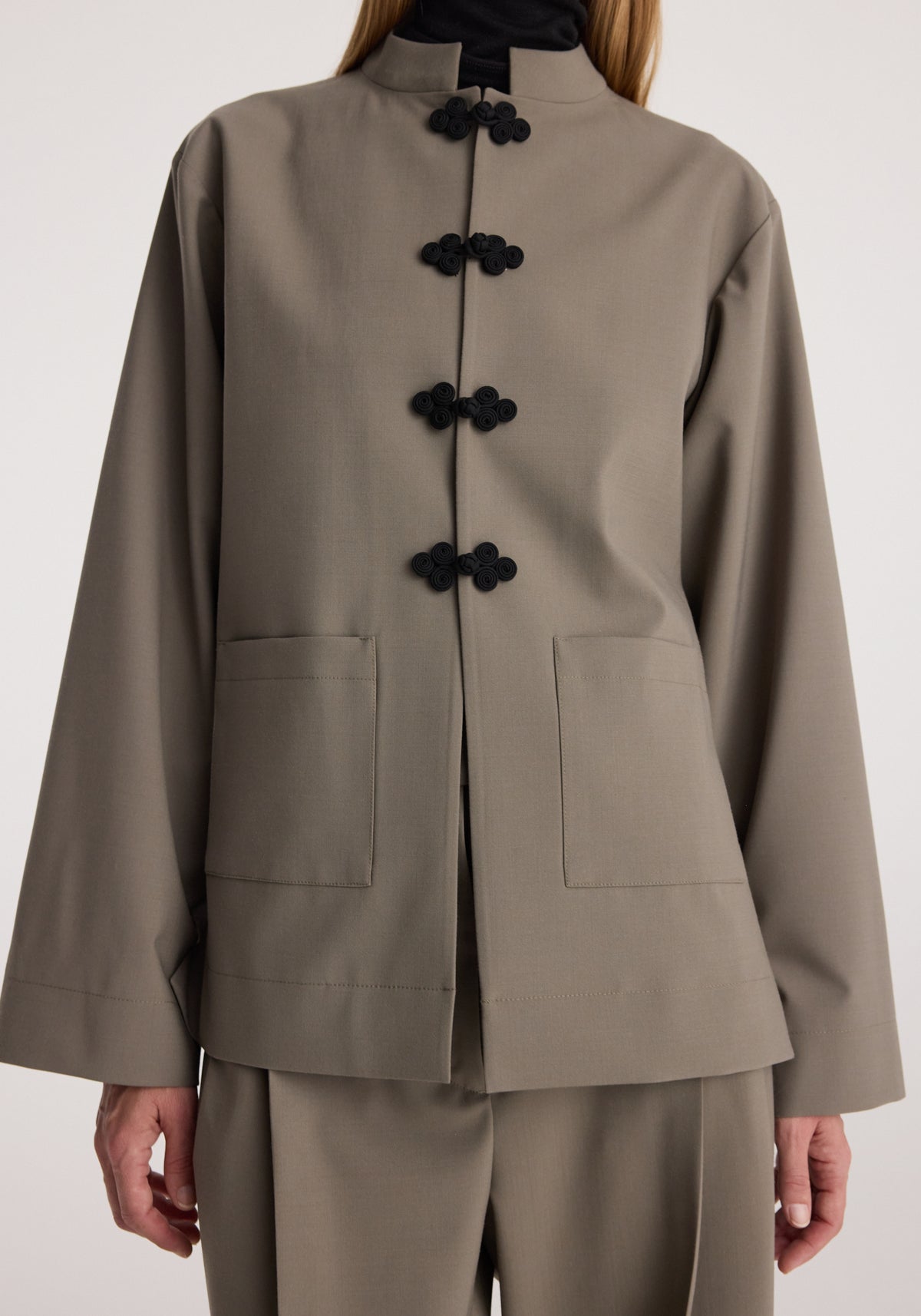 Pankou closure jacket | taupe shown on a model, featuring a high collar, black knotted closures, long sleeves, front pockets, and a relaxed tailored fit.