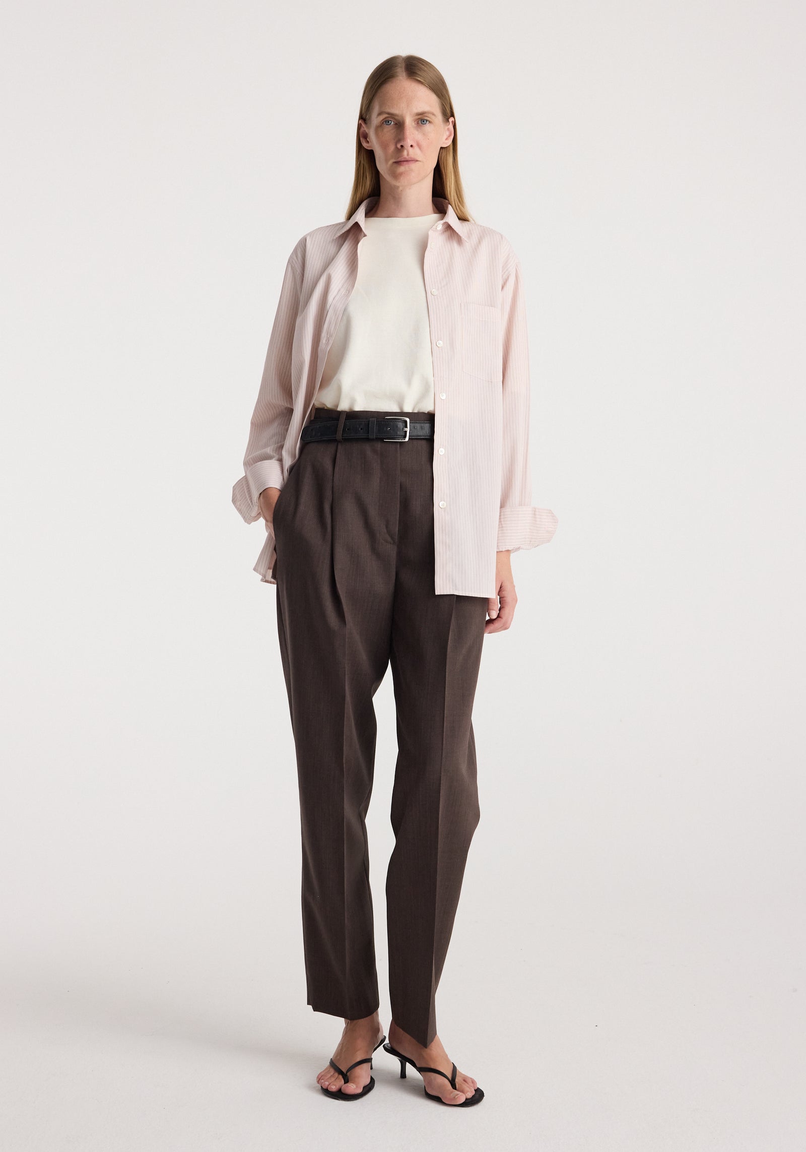 Woman wearing a Classic fit striped shirt | rose stripe layered over a white top, paired with dark high-waisted tailored pants and black heeled sandals, standing against a plain white background.