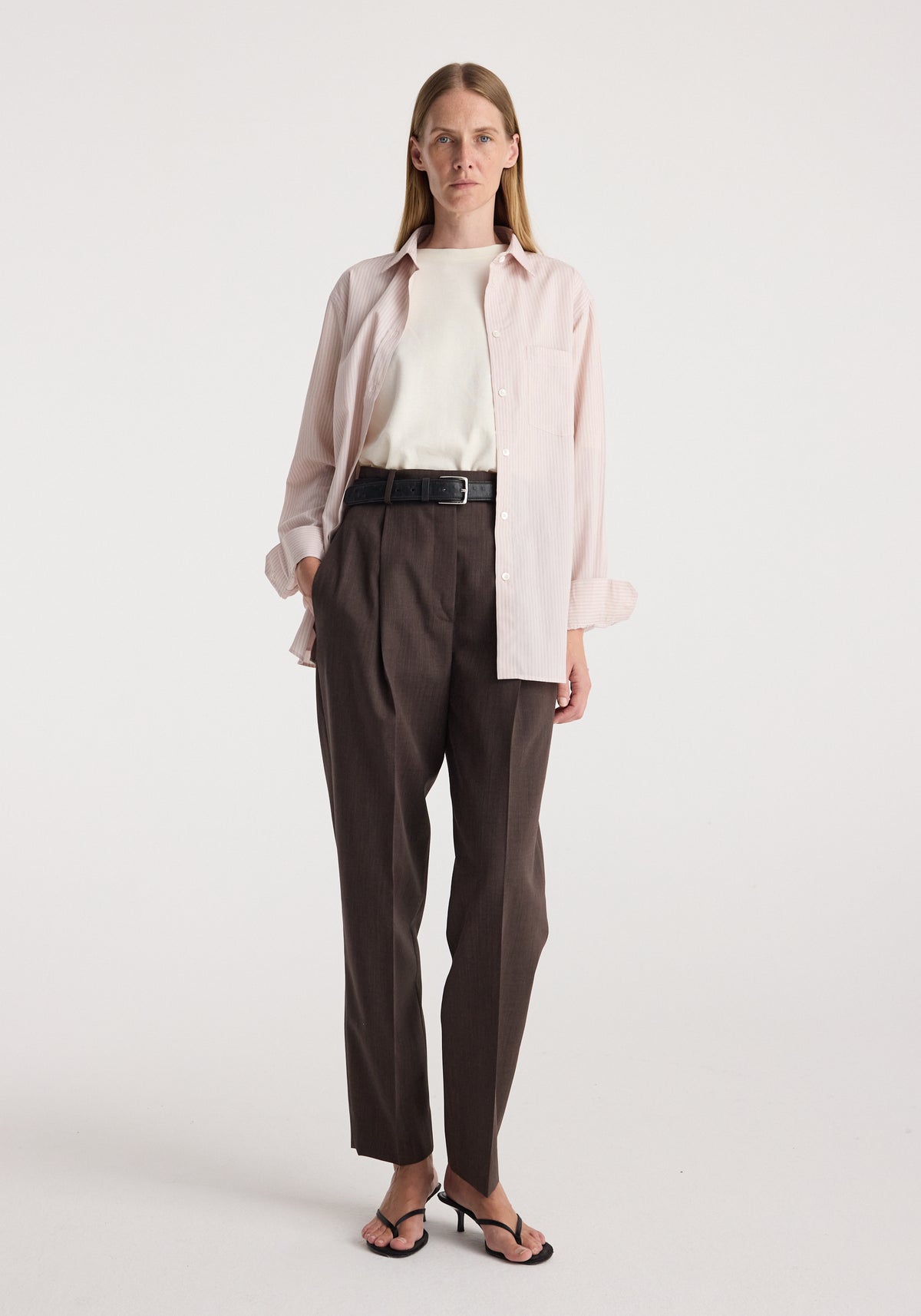 Woman wearing a Classic fit striped shirt | rose stripe layered over a white top, paired with dark high-waisted tailored pants and black heeled sandals, standing against a plain white background.