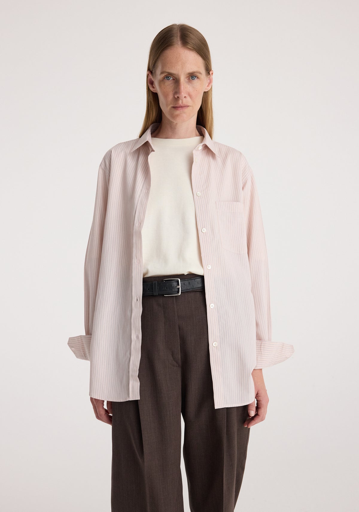 Woman wearing a Classic fit striped shirt | rose stripe, light pink with subtle vertical stripes, styled open over a white top, paired with dark brown trousers and a black belt.