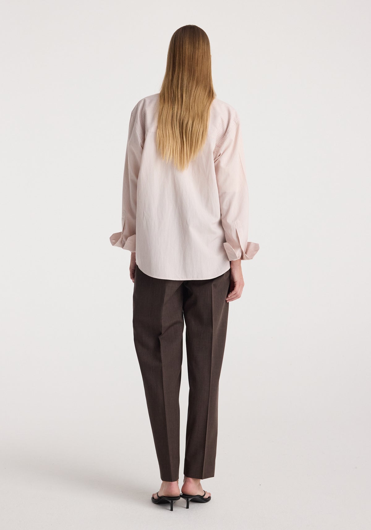 Rear view of a person wearing a Classic fit striped shirt | rose stripe in light pink with long sleeves, paired with dark straight-leg pants and black heeled sandals.