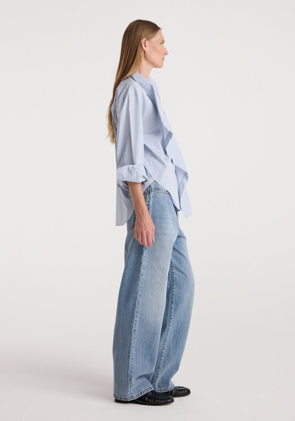 Side view of model wearing Draped collar shirt | blue stripe, a light blue striped shirt with draped collar and flared cuffs, paired with wide leg jeans