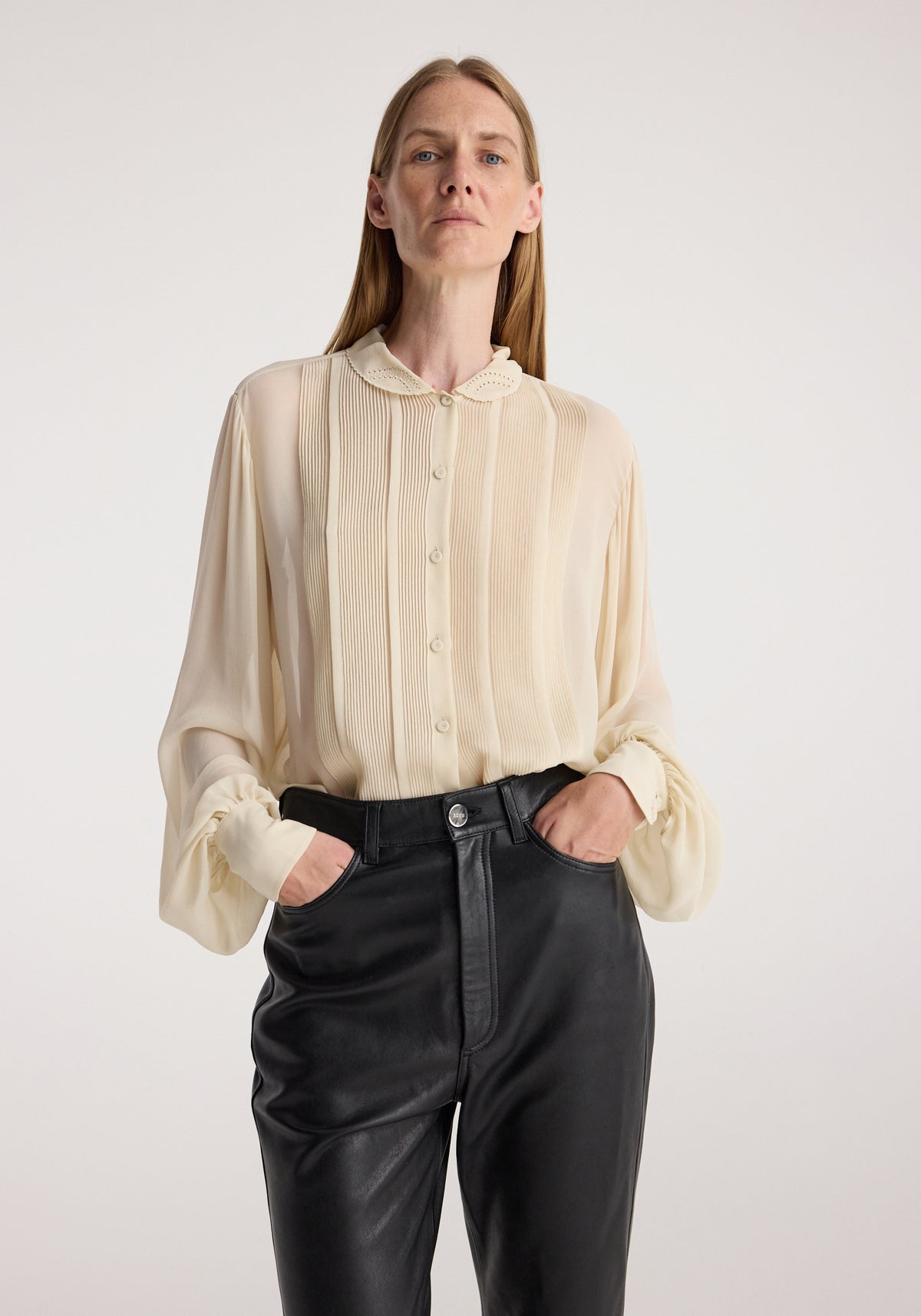 Woman wearing a cream pleated blouse with button-up front and scalloped collar tucked into black leather pants, styled with hands in pockets, Romantic top | cream.