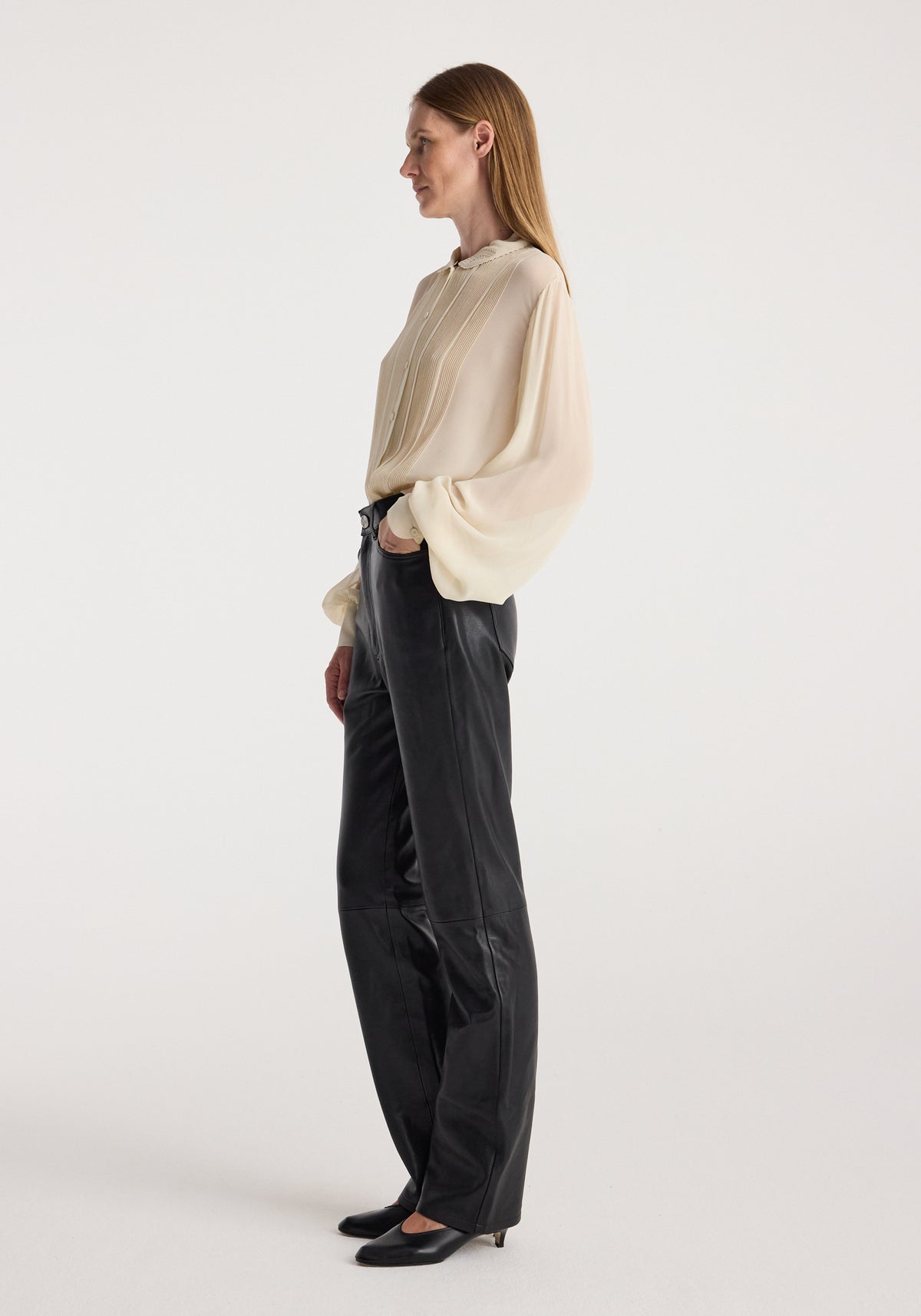 Woman wearing a cream romantic top with long balloon sleeves and pleated front paired with black leather pants and black heels, standing in profile against a white background.