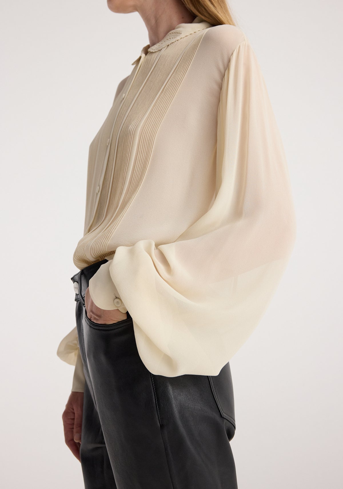 Woman wearing a cream-colored Romantic top | cream with sheer, billowy long sleeves and pintuck detailing on the front, paired with black leather pants.